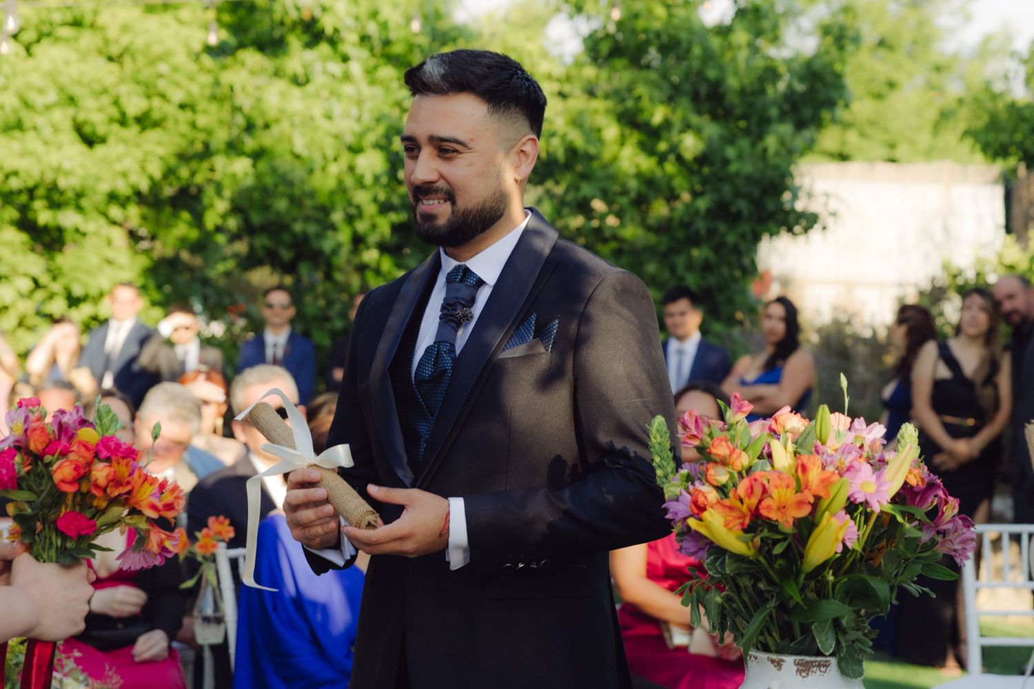 Groom in navy suit smiling at outdoor wedding ceremony with colorful floral arrangements.