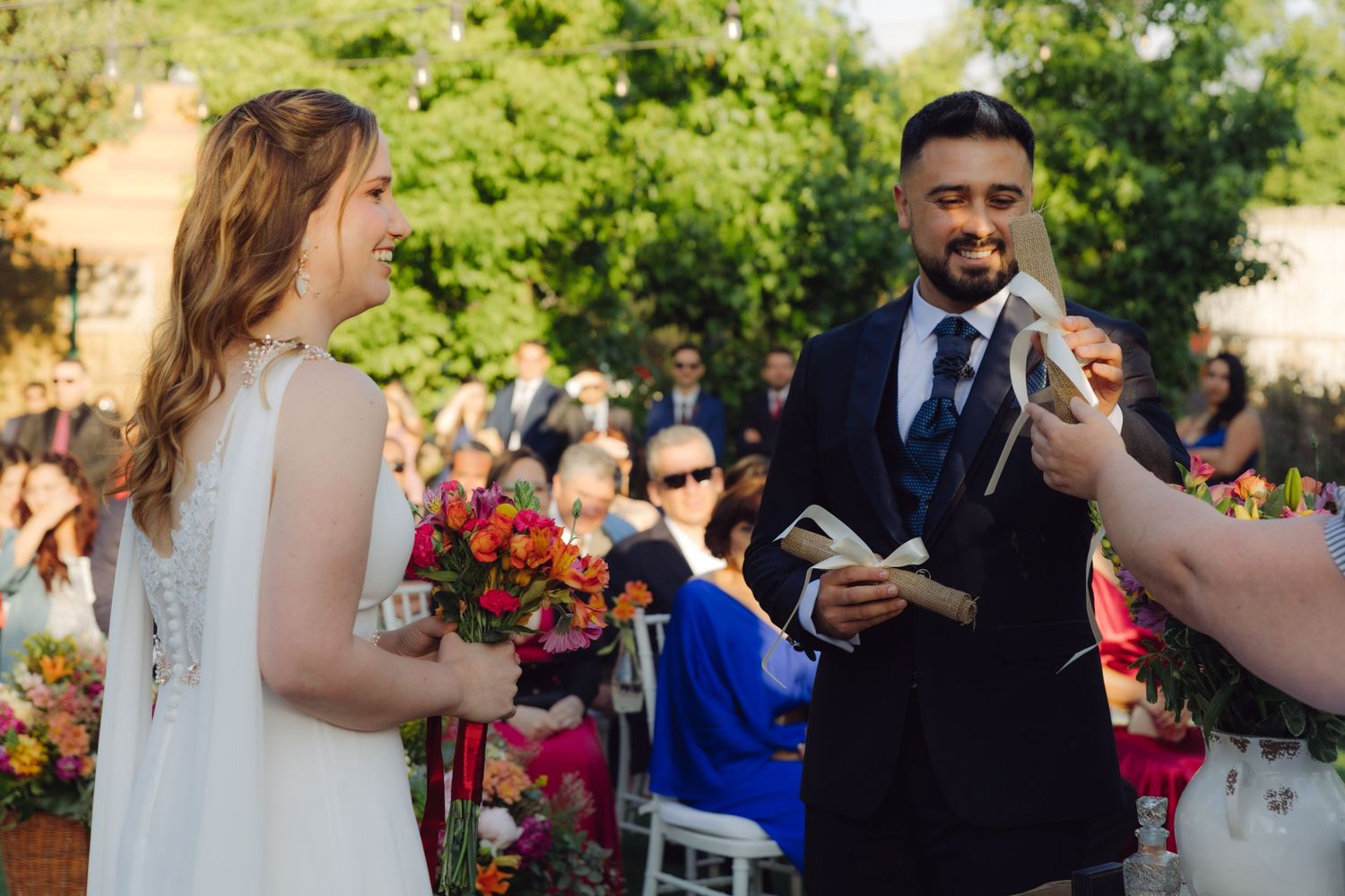 Groom smiling at bride during outdoor wedding ceremony with colorful bouquets and guests.