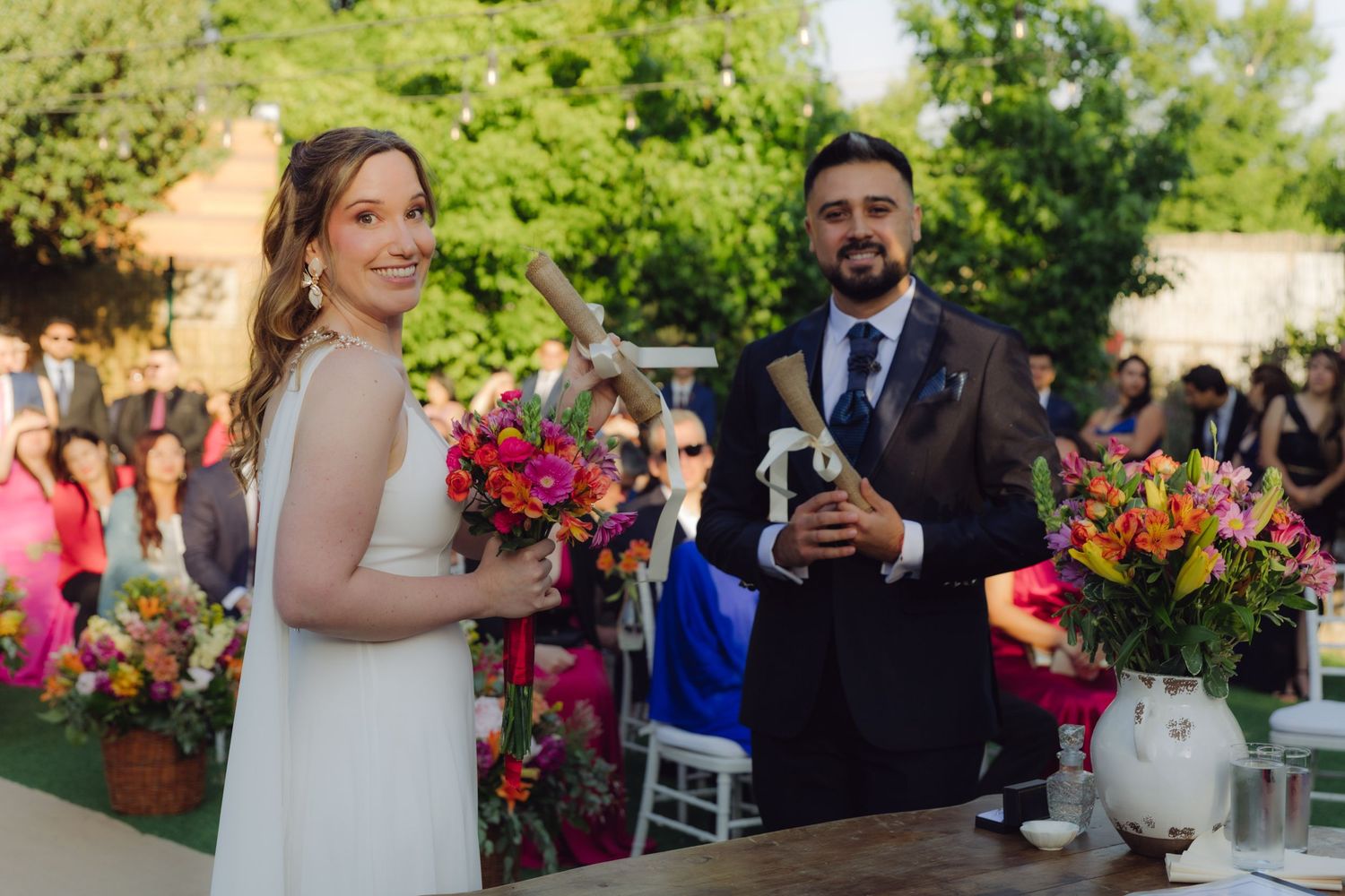 Bride and groom smiling together at outdoor wedding ceremony holding bouquet and scroll.