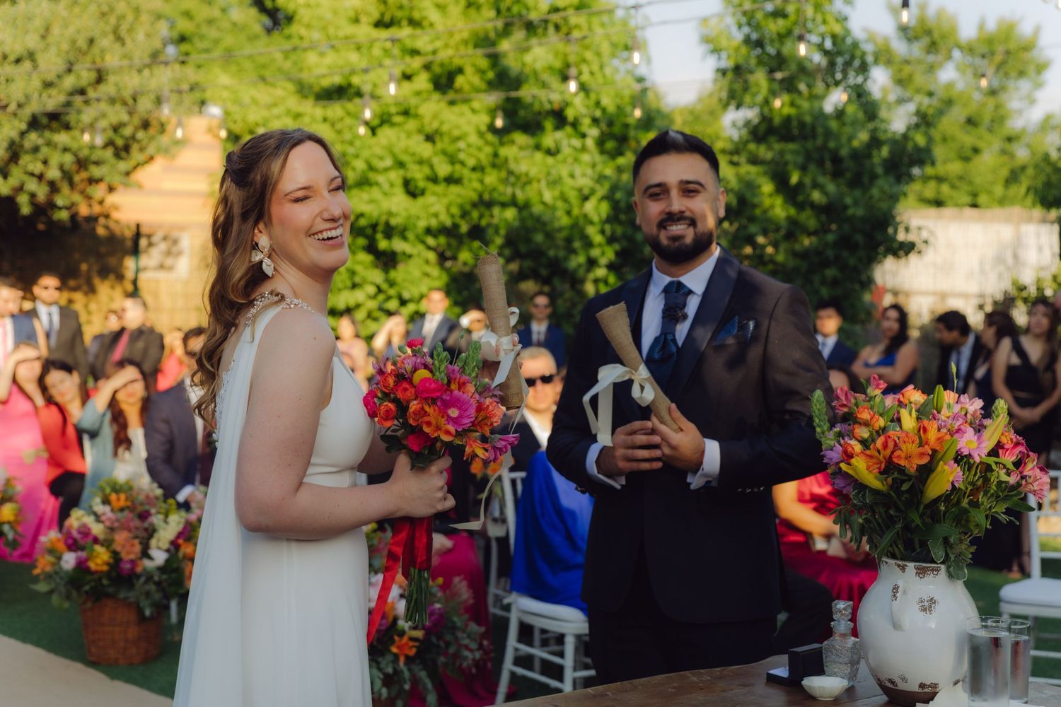 Happy bride and groom laughing together at outdoor garden wedding ceremony with colorful flowers.
