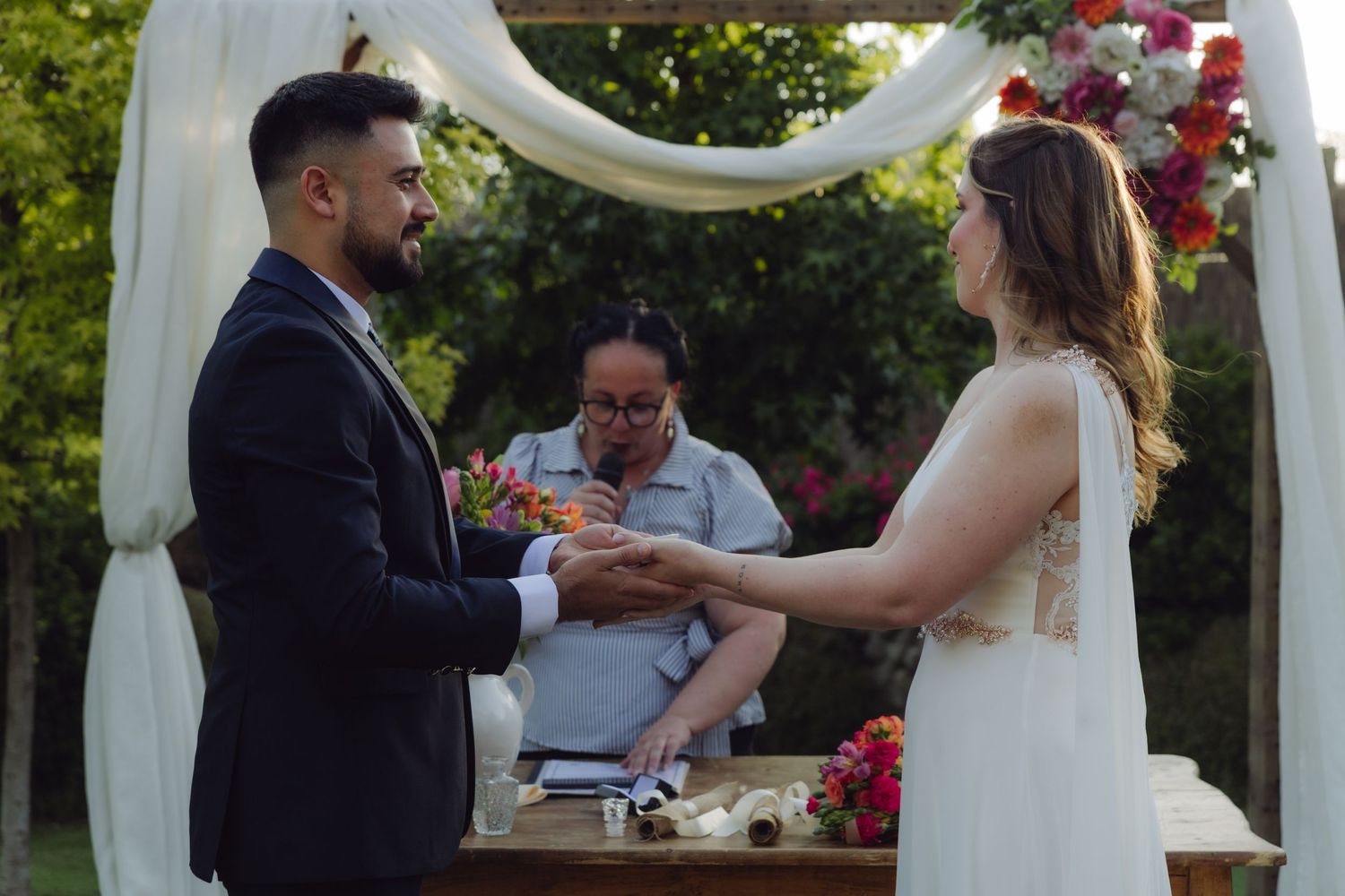 Groom placing ring on bride's hand at outdoor garden wedding ceremony under floral arch.