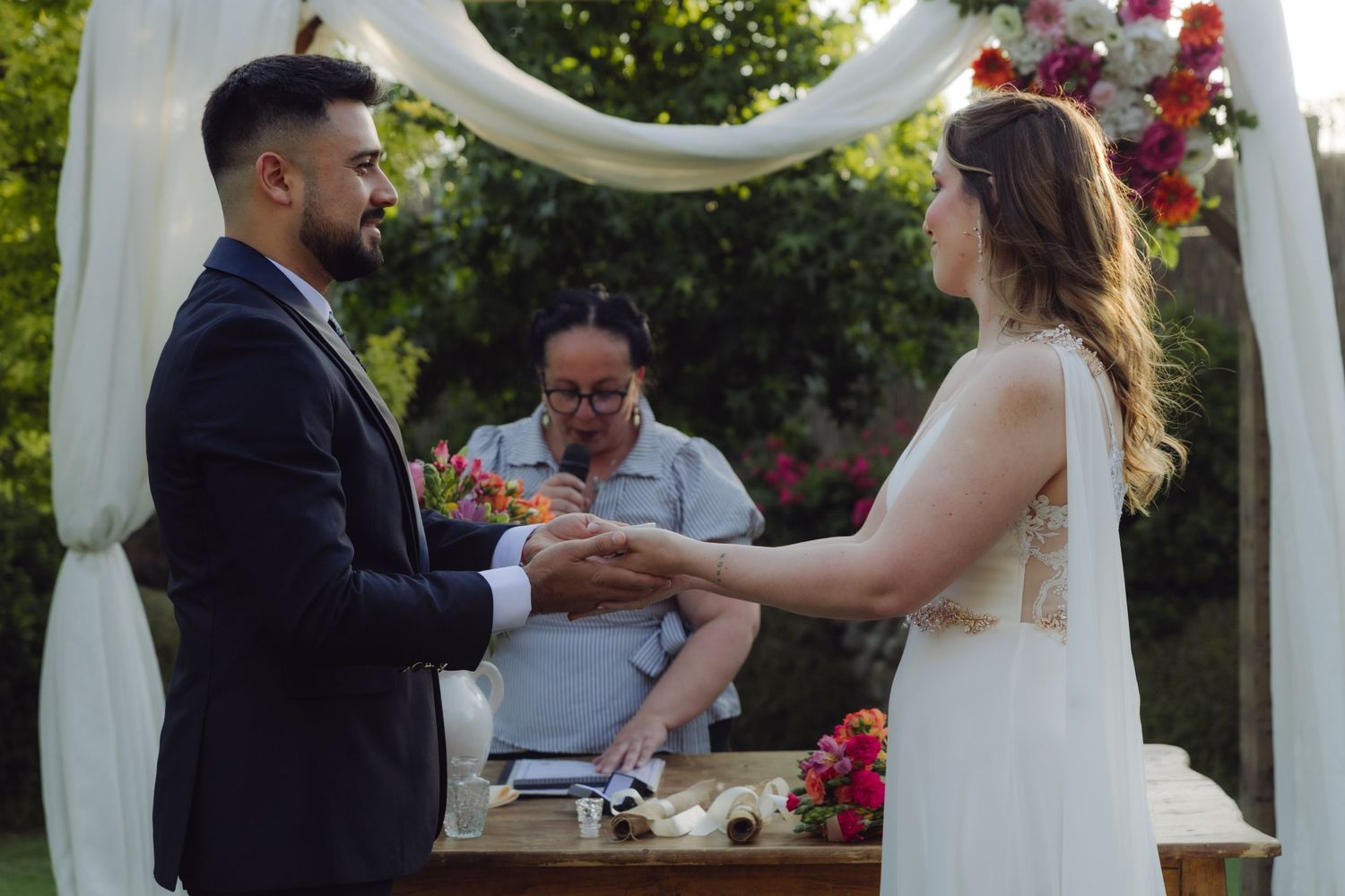 Groom exchanging rings with bride at outdoor garden wedding ceremony under decorated arch.