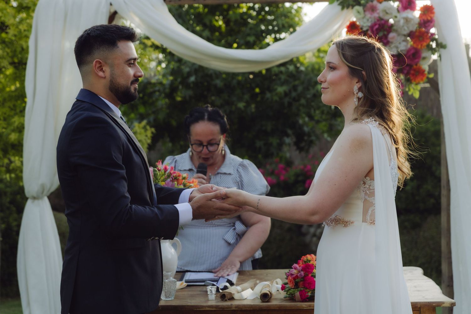 Groom holding bride's hand during ring exchange at outdoor garden wedding ceremony.