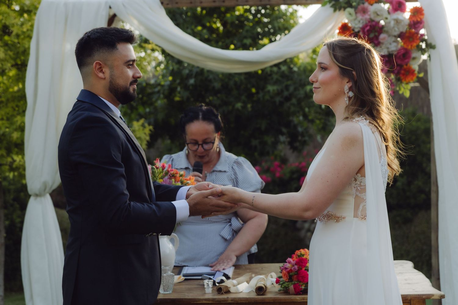 Couple exchanging rings at outdoor garden wedding ceremony under white draped floral arch.