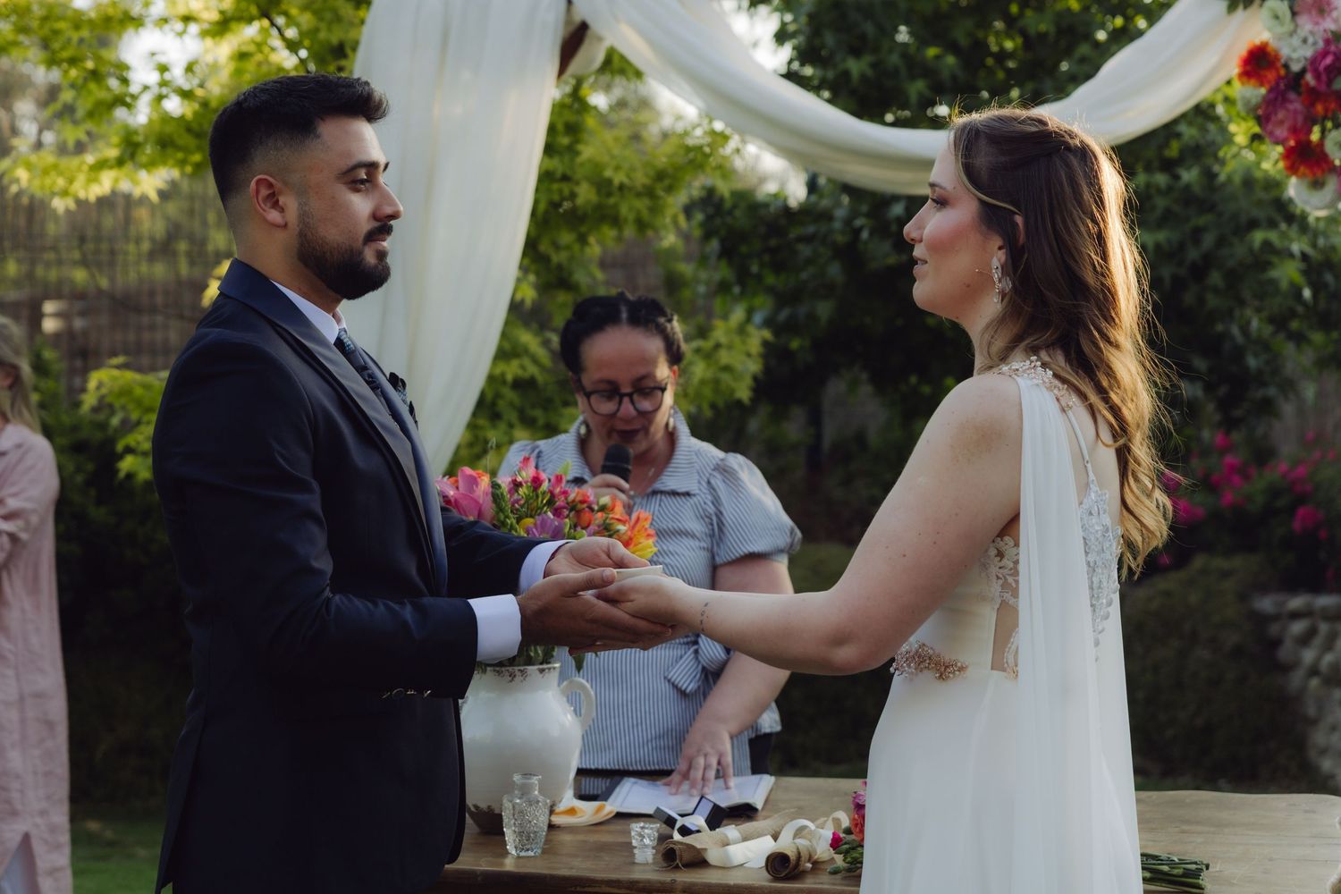 Bride and groom exchange rings during an outdoor wedding ceremony beneath a floral arch.