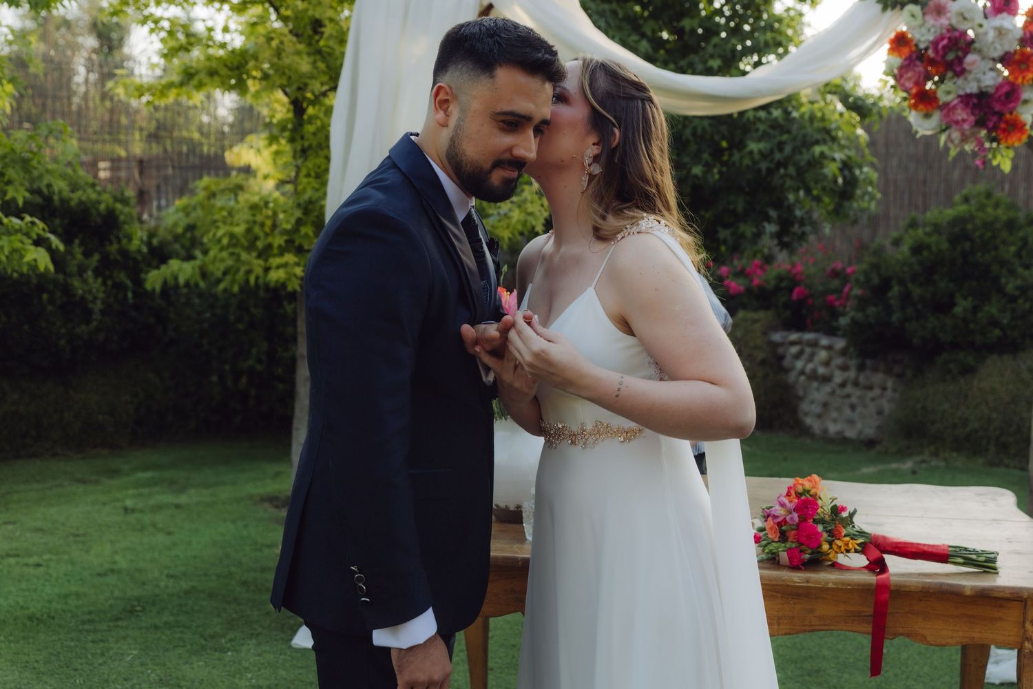 Newlyweds share their first kiss beneath a white draped arch at an outdoor wedding.