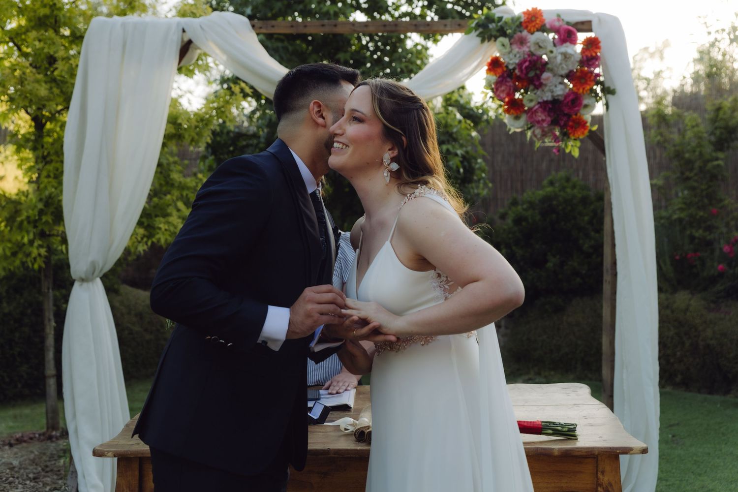 Bride and groom kiss passionately under a colorful floral arch at their garden wedding.