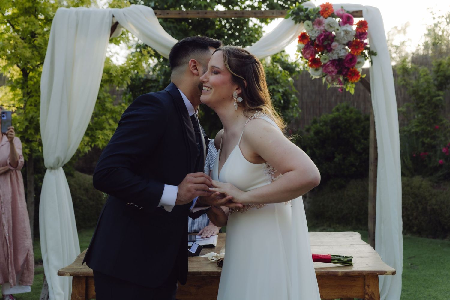 Happy newlyweds share a romantic kiss beneath a white and floral wedding arch outdoors.