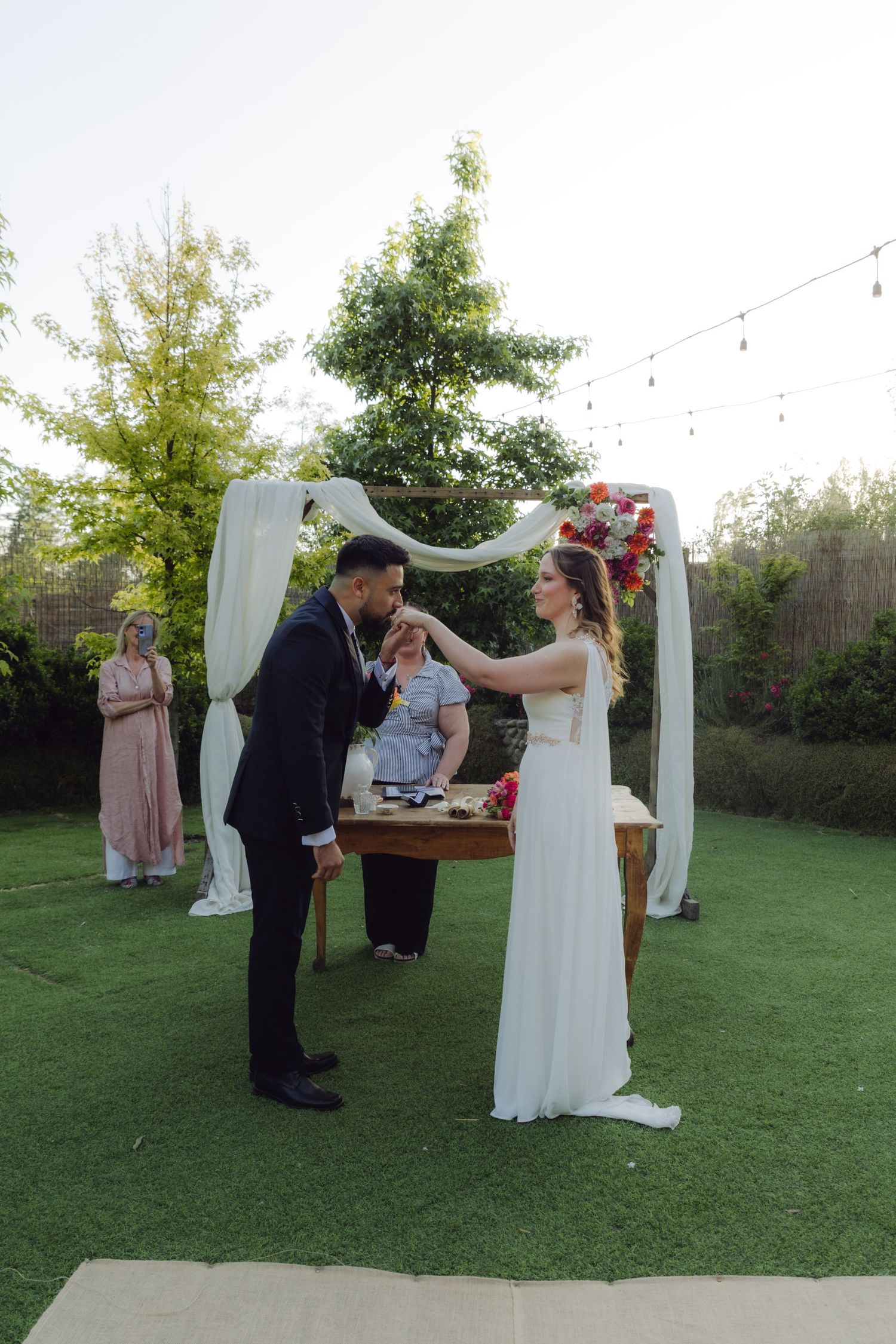 Groom kisses bride's hand during an elegant outdoor garden wedding ceremony at dusk.