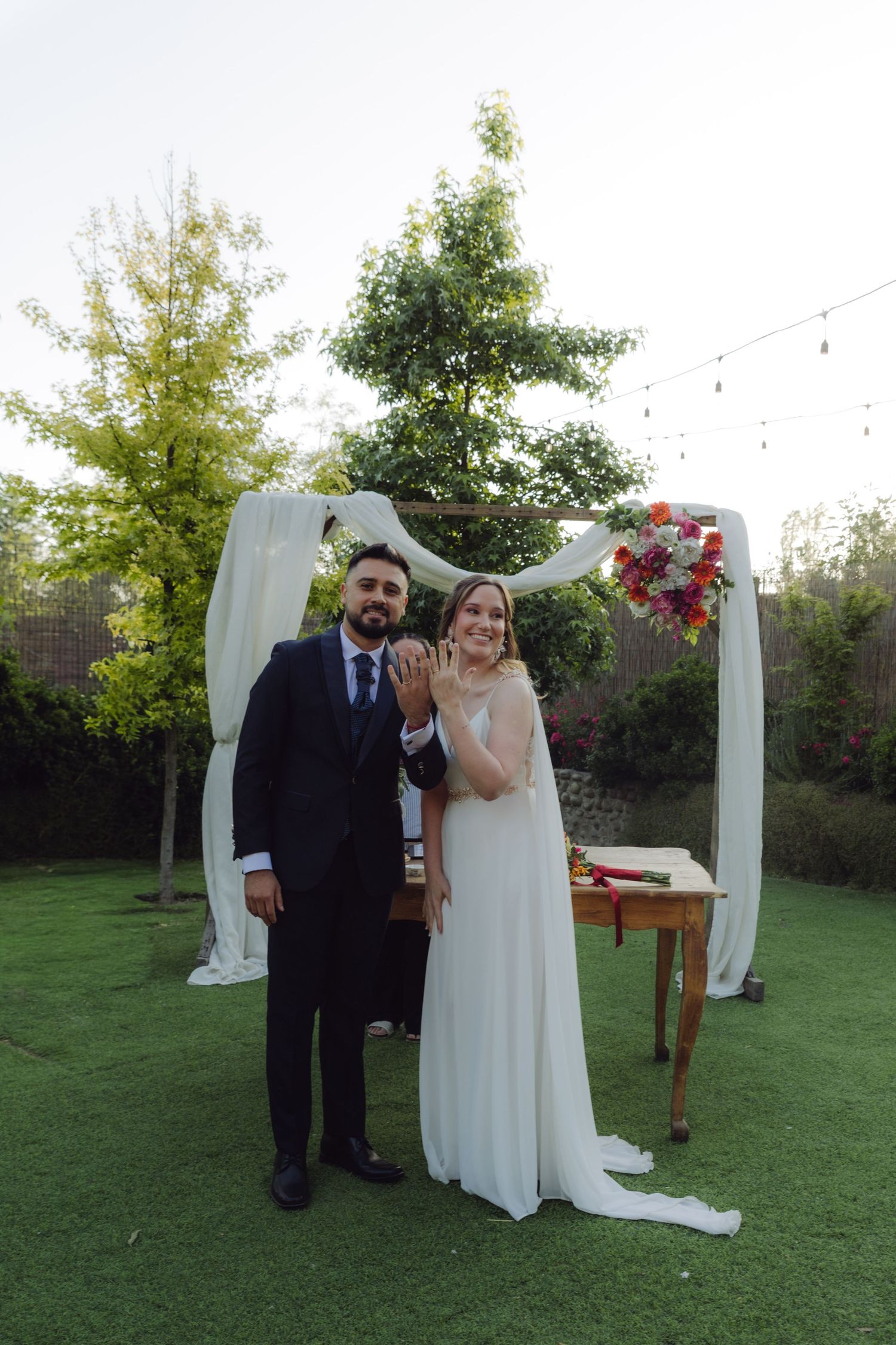 Newlyweds proudly display their wedding rings while posing under a floral arch outdoors.