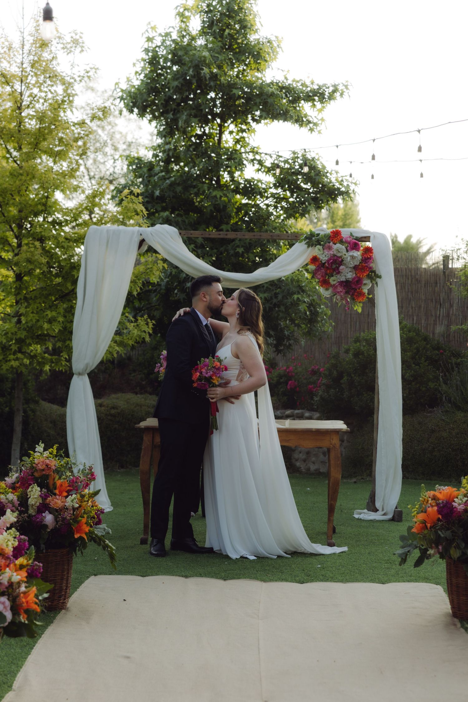 Bride and groom share their first kiss under a floral arch at an outdoor wedding ceremony.