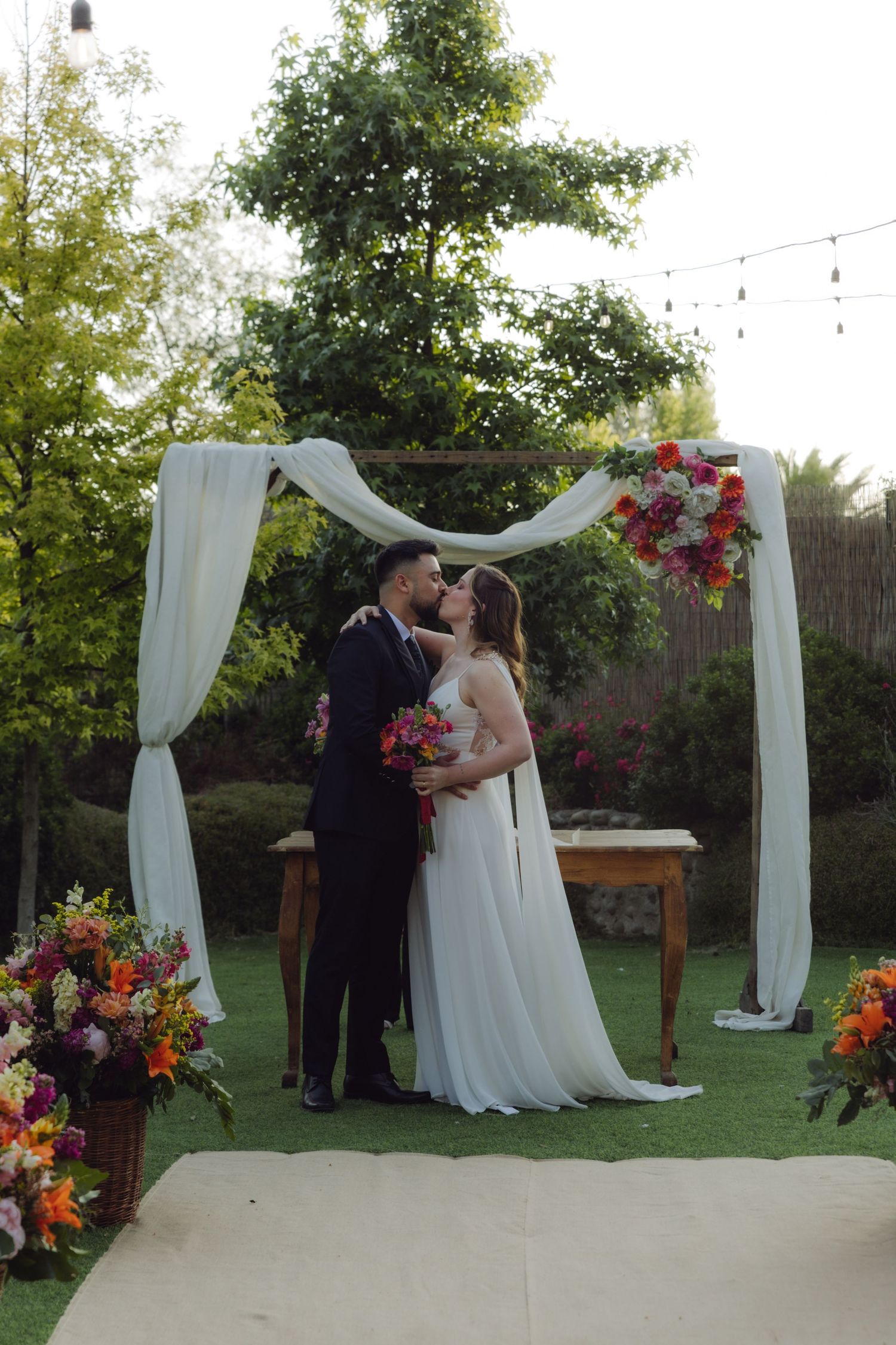Newlyweds kiss under a white draped floral arch surrounded by colorful flowers at their wedding.