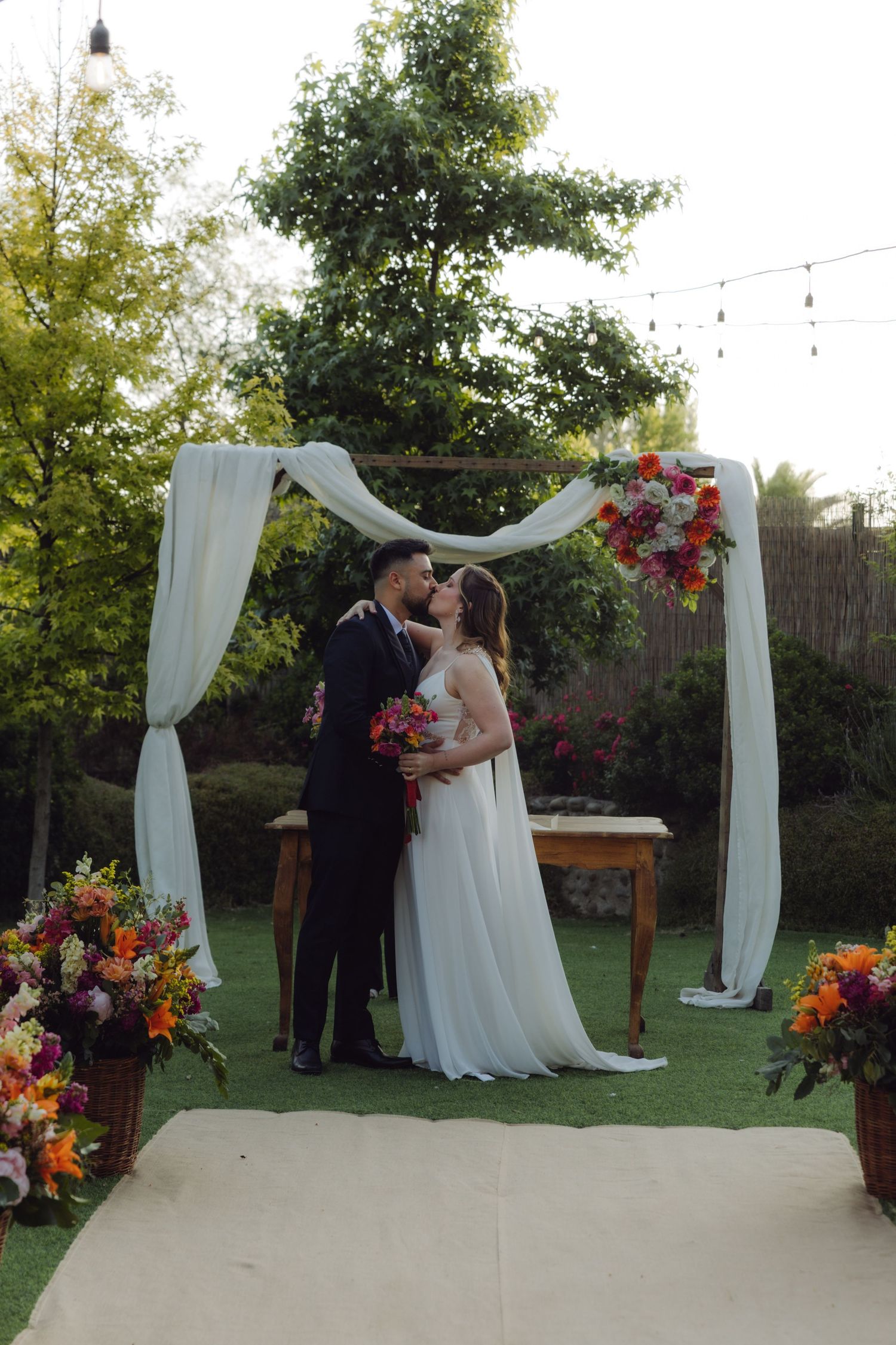 Bride holding a vibrant bouquet kisses groom under a decorated arch at an outdoor wedding.
