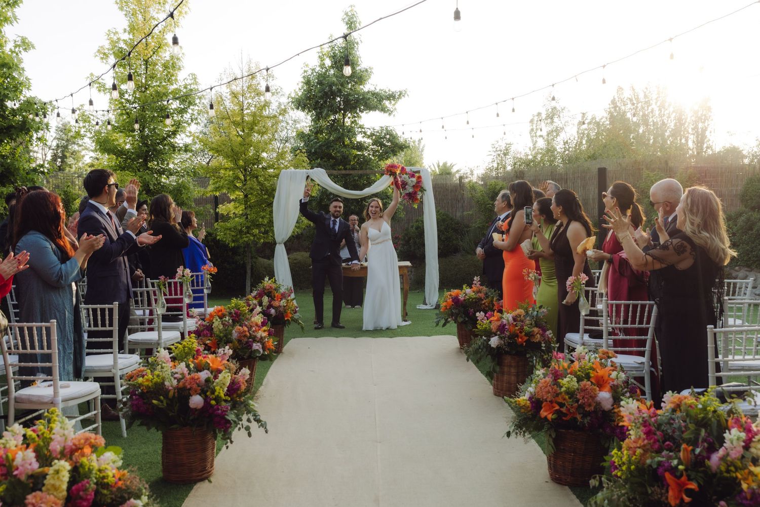 Newlyweds walk down the aisle at an outdoor wedding ceremony with colorful floral arrangements.