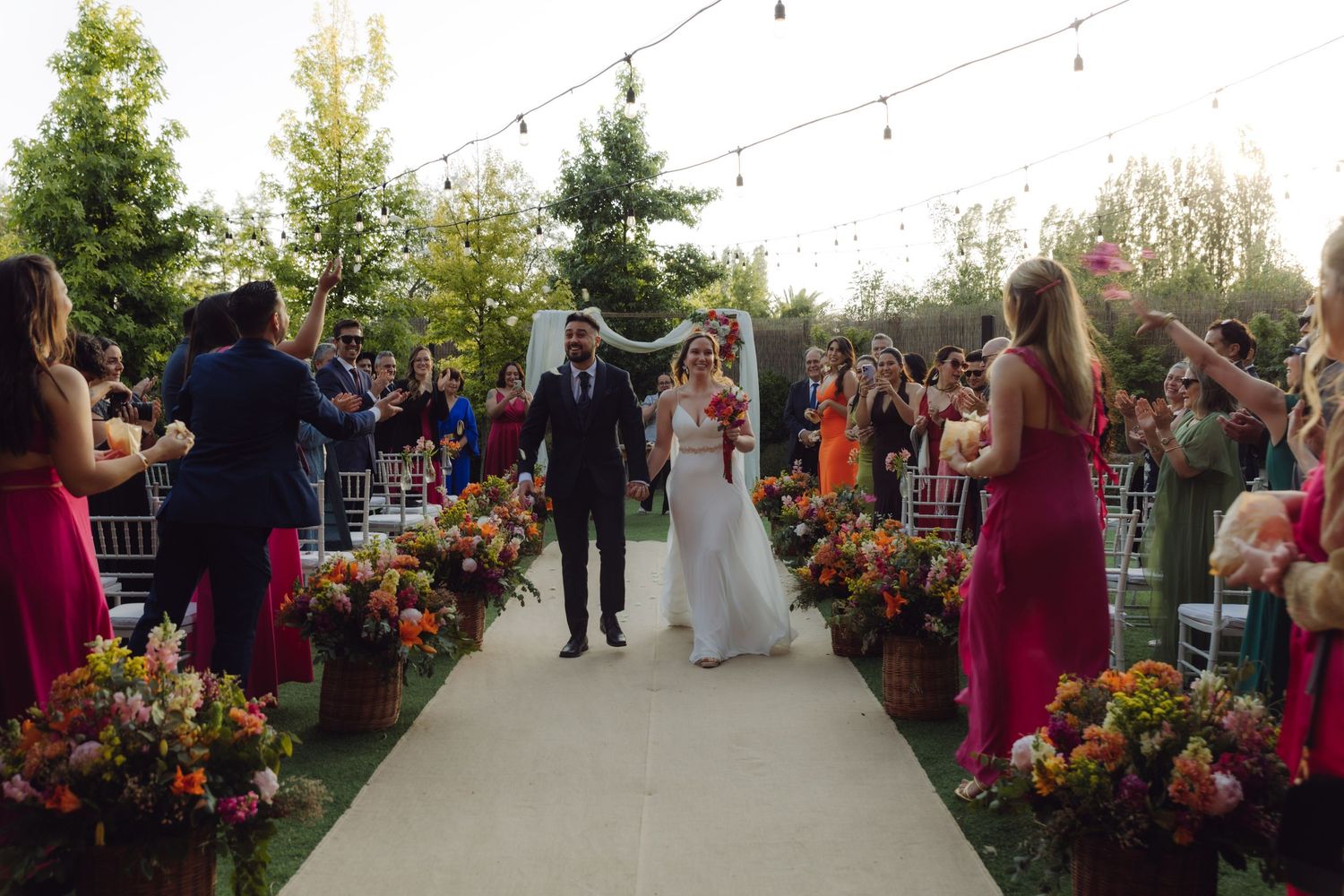 Bride and groom walk down the flower-lined aisle at a festive outdoor wedding ceremony.