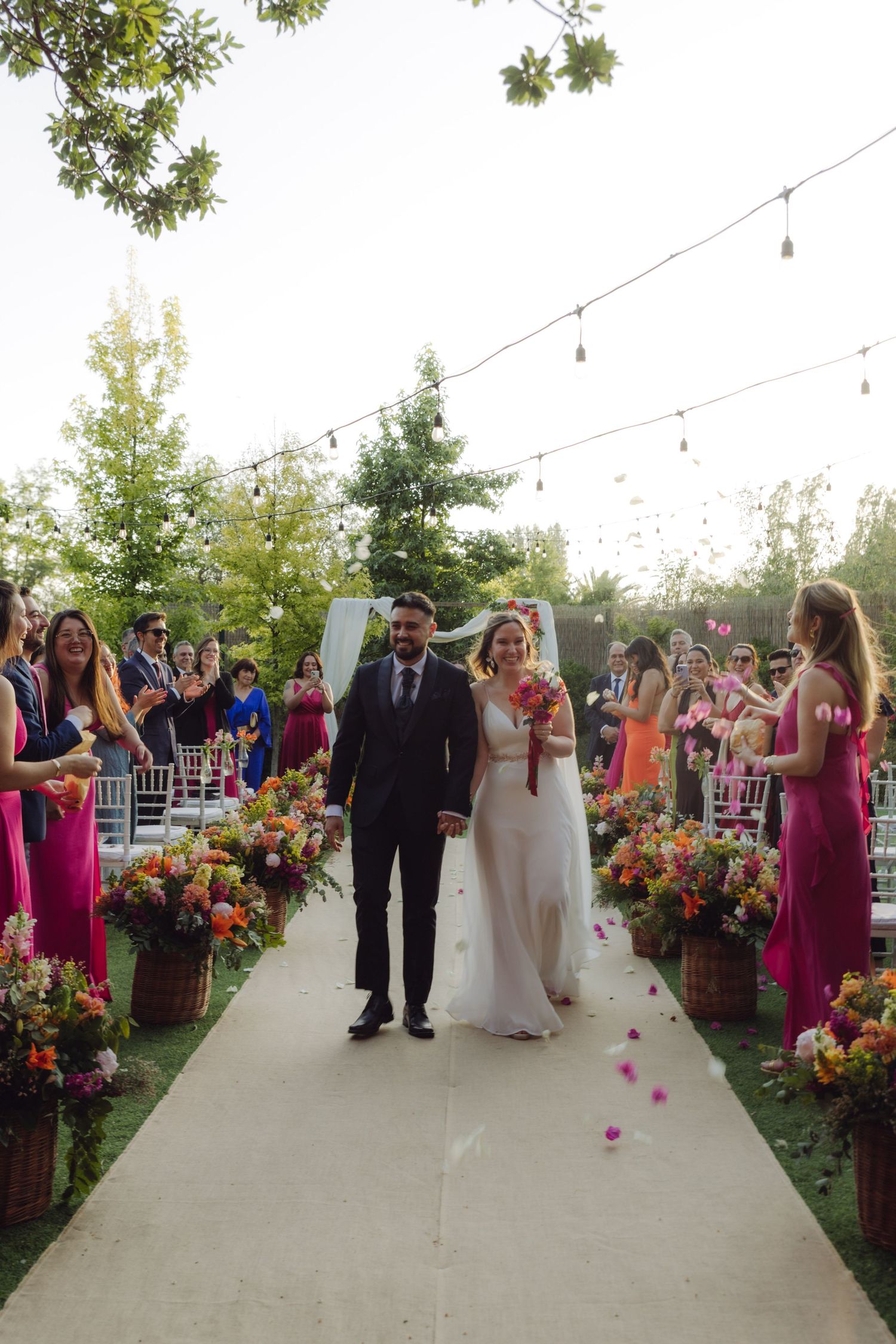 Newlyweds walk down a petal-strewn aisle at an outdoor wedding with string lights above.