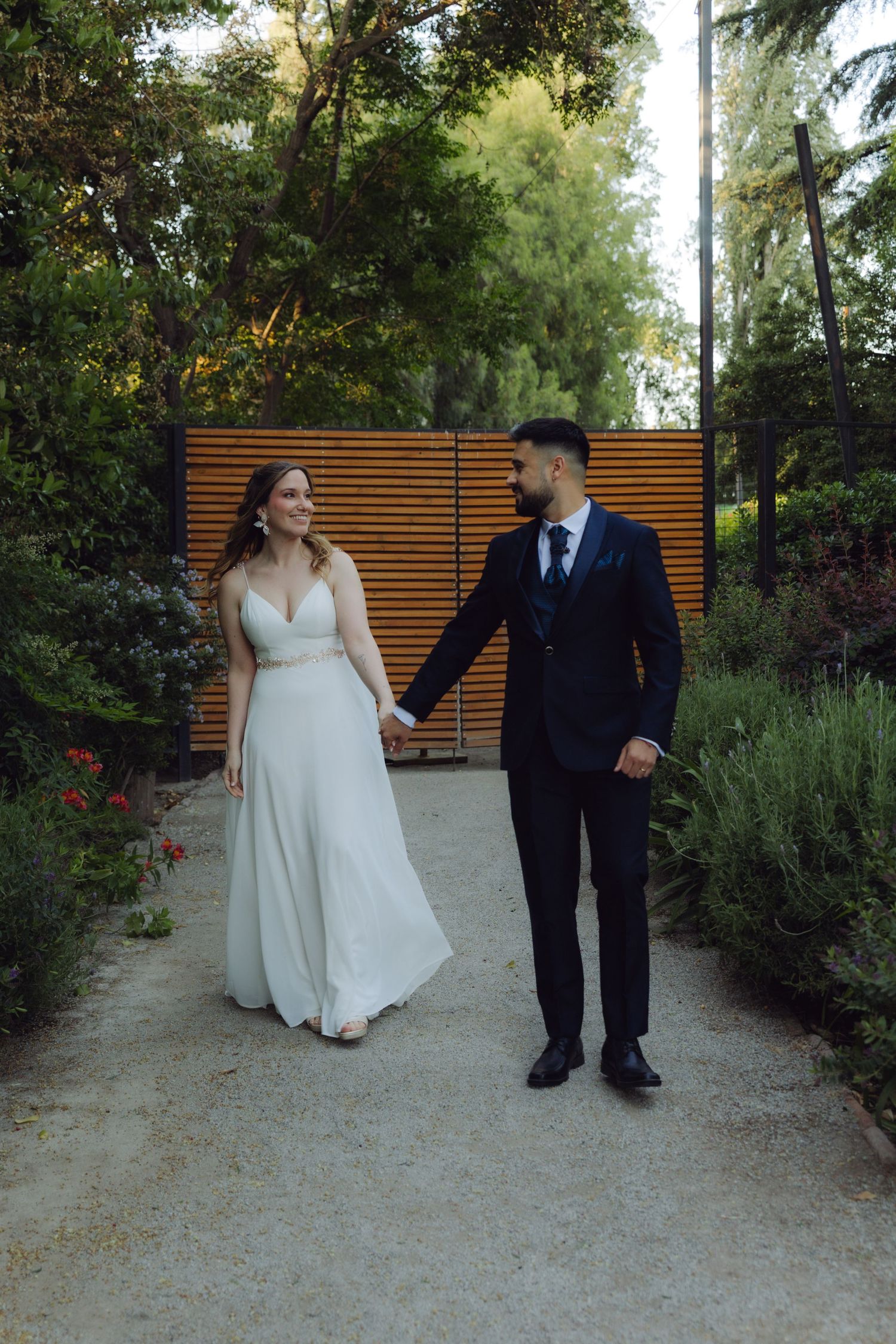 Wedding couple walks hand in hand through a lush garden pathway at dusk.