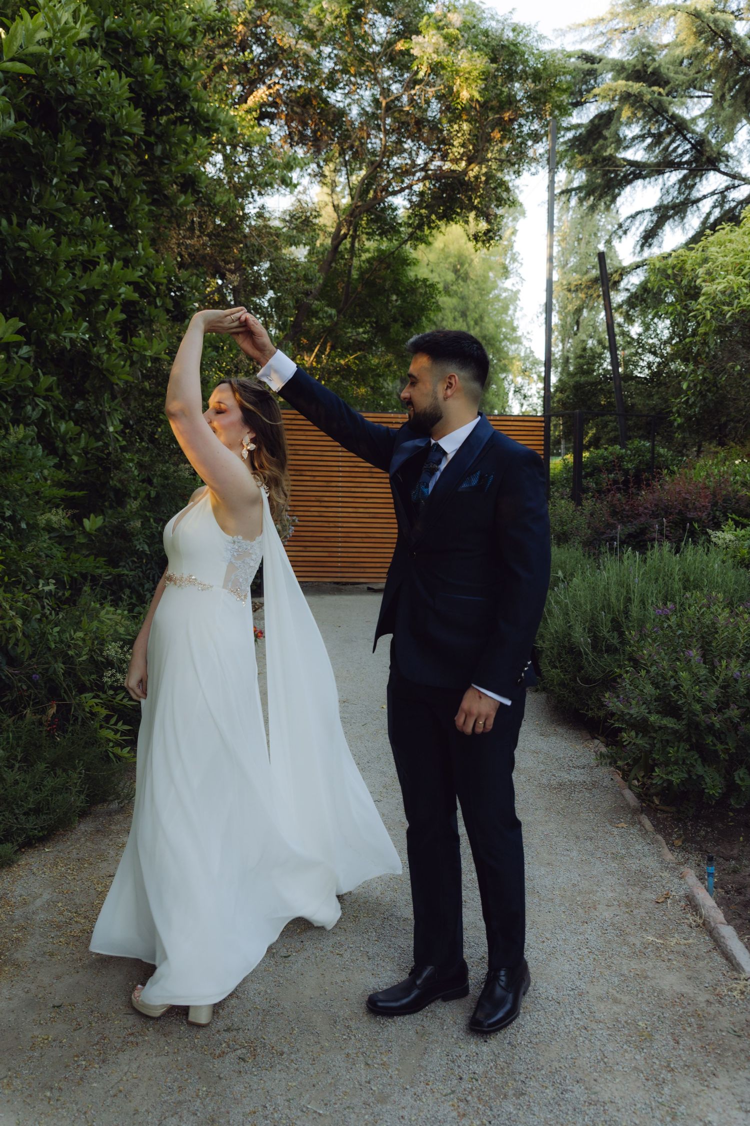 Groom twirls bride in white gown on a garden path surrounded by greenery.