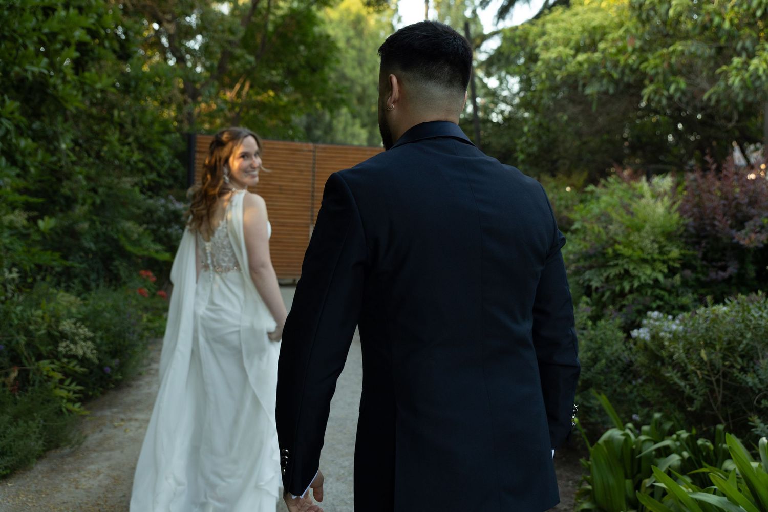 Bride in backless white gown leads groom along a tree-lined garden path.