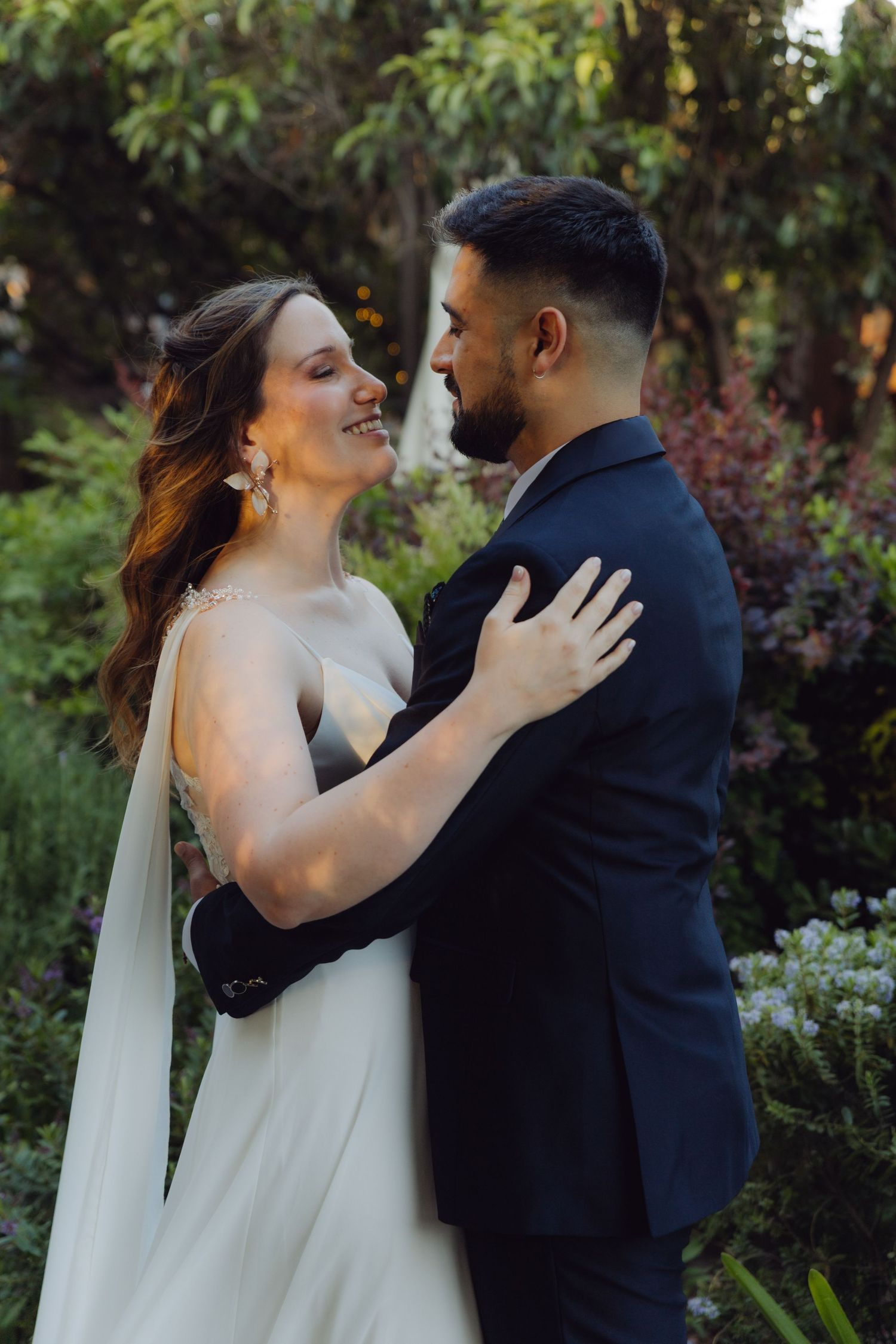 Smiling bride in white dress embraces groom in navy suit in a garden setting.