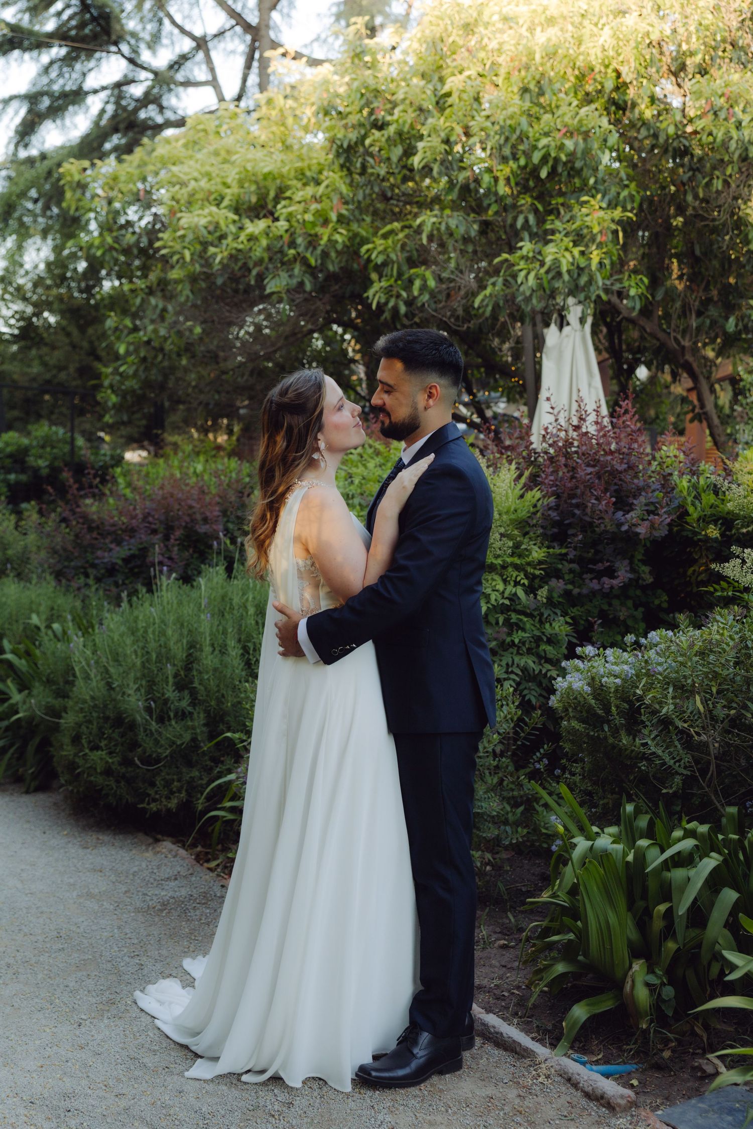 Newlywed couple shares a tender embrace on a garden path surrounded by lush plants.