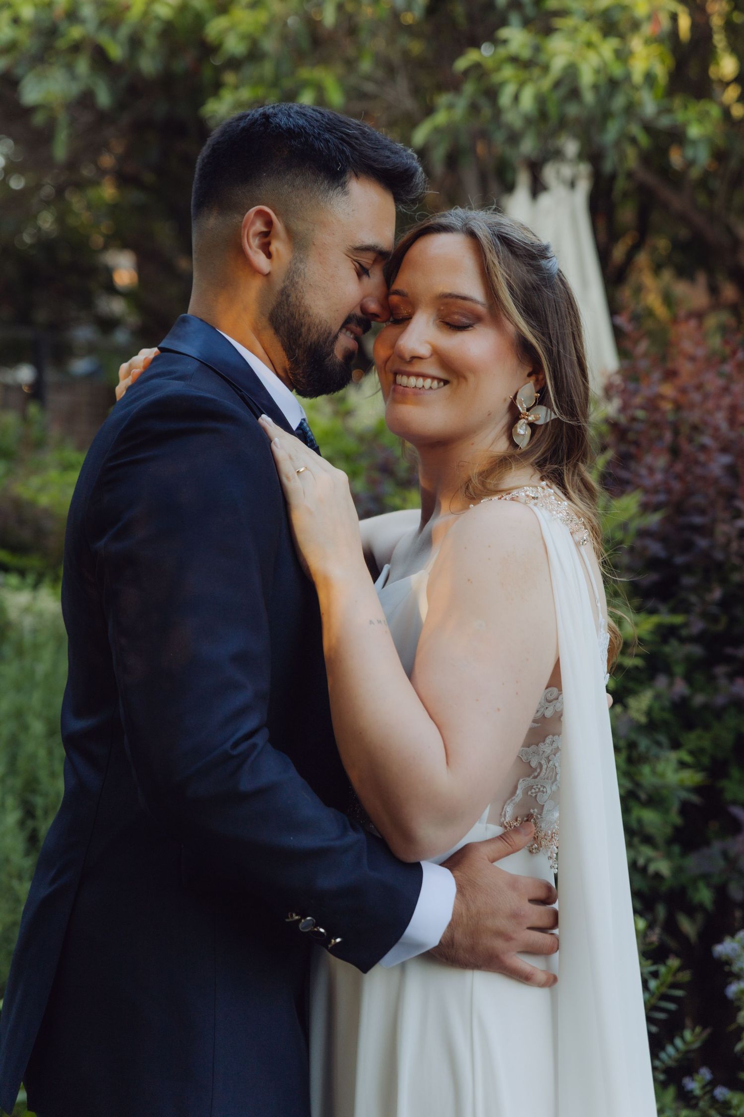 Groom nuzzles smiling bride wearing statement earrings in a romantic garden portrait.
