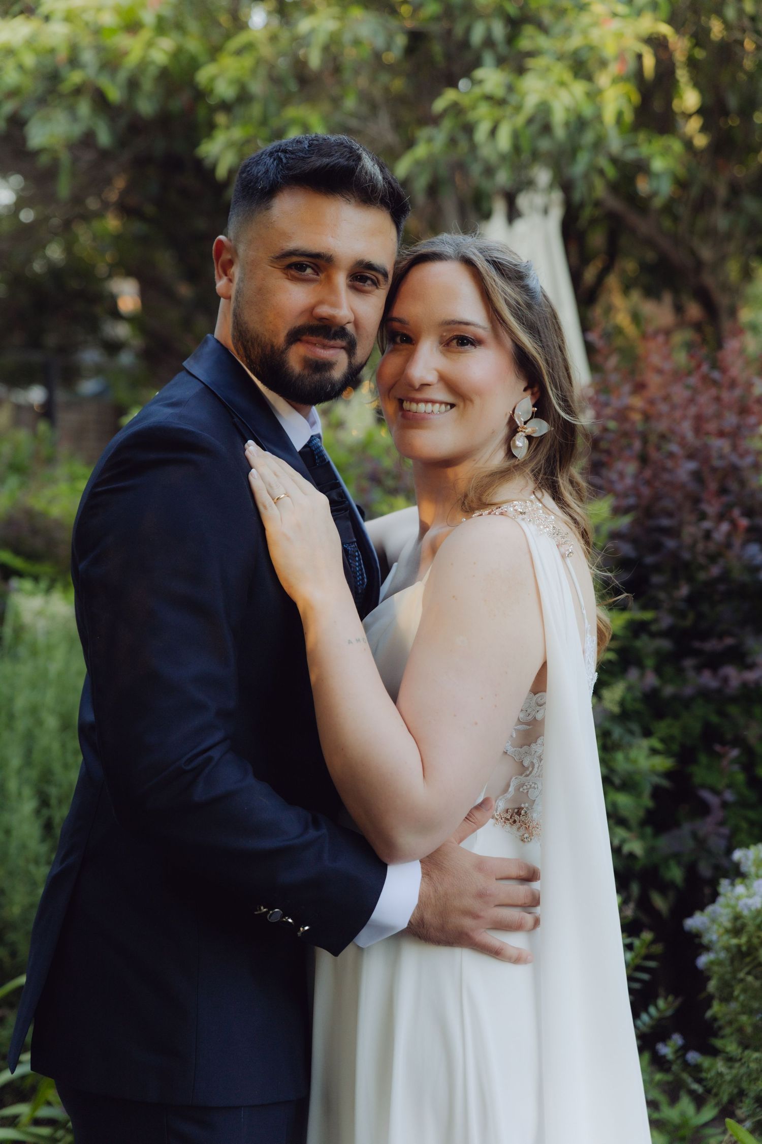 Newlywed couple embraces and smiles at camera in a lush outdoor garden setting.