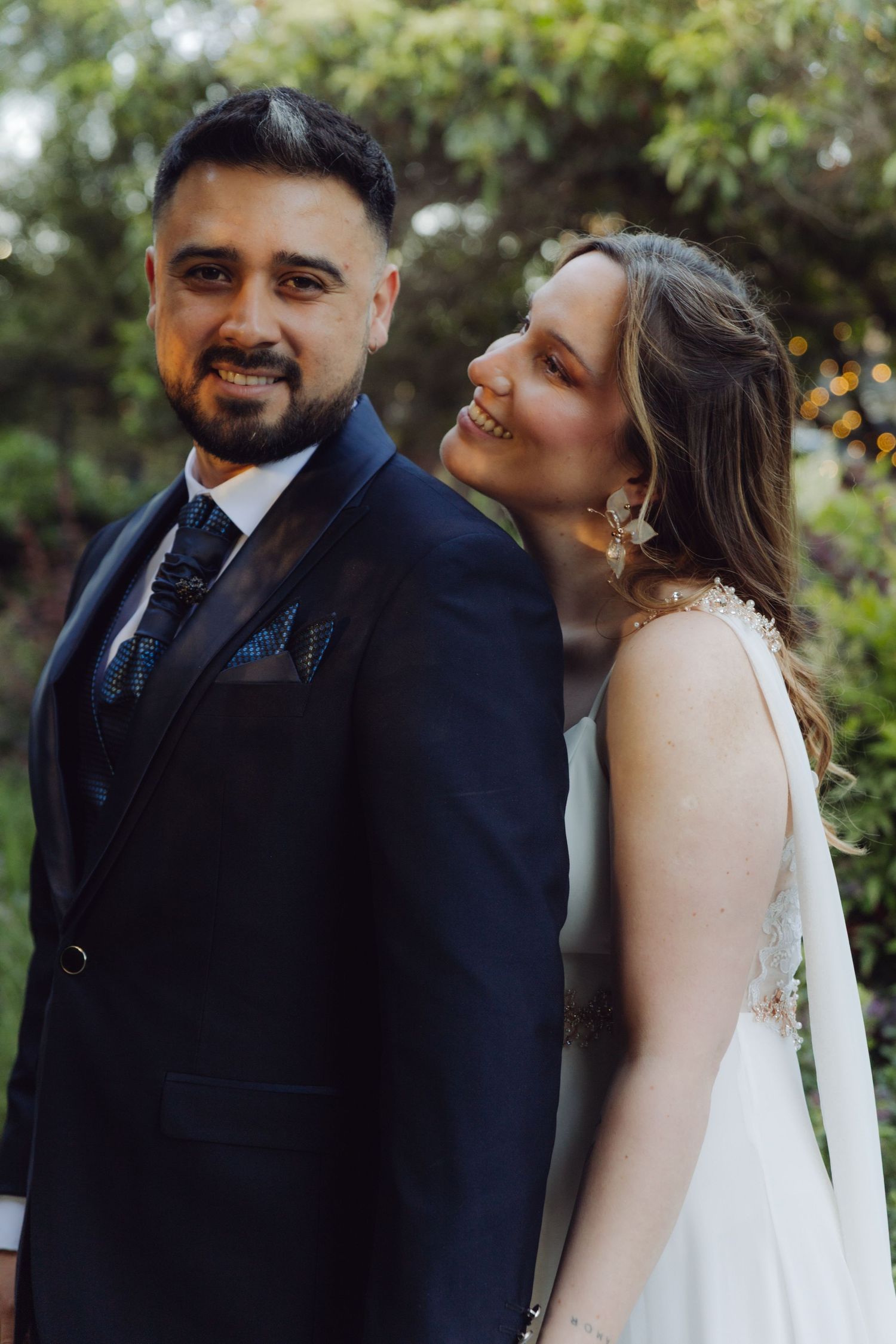 Groom smiles at camera while bride leans in close laughing in garden portrait.