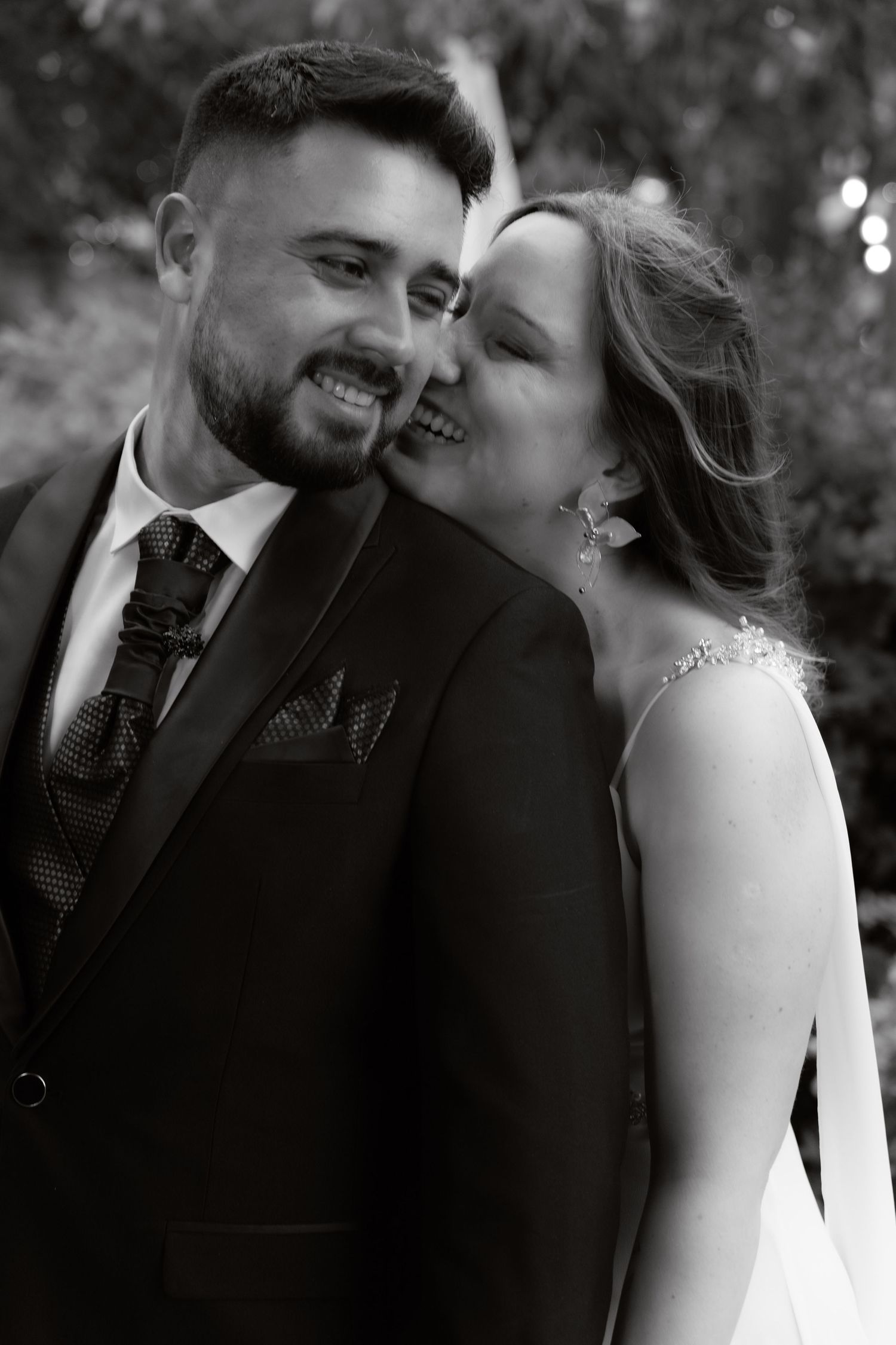 Black and white photo of a smiling groom in a suit as the bride kisses his cheek.