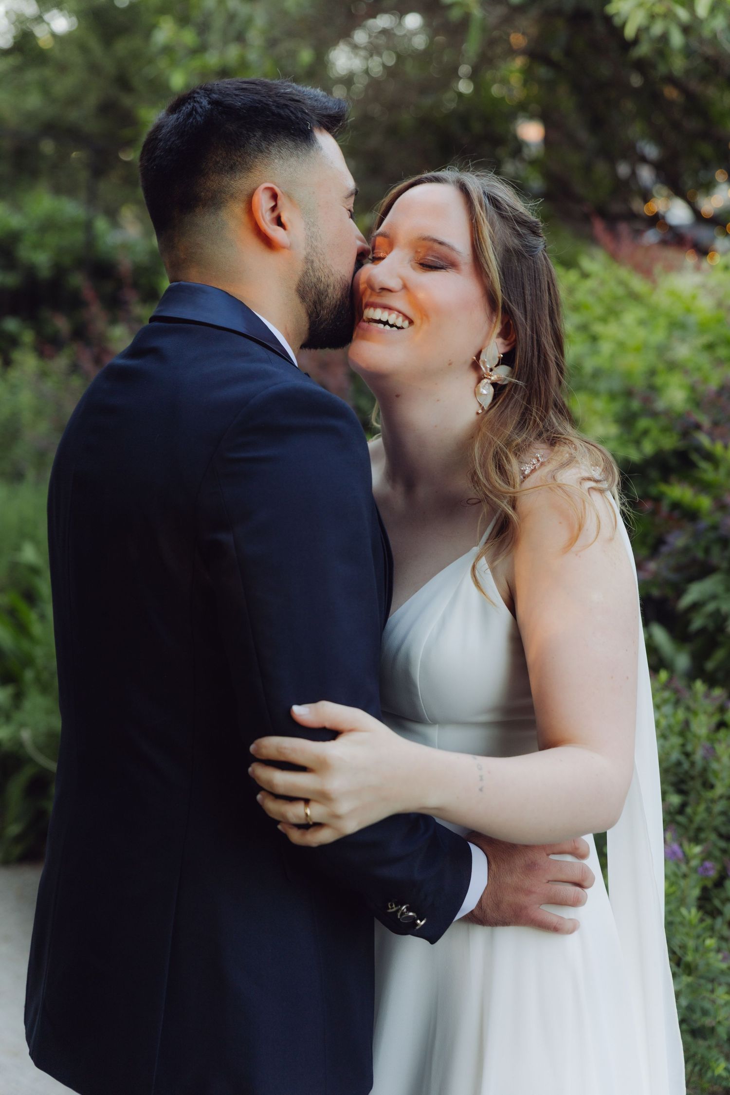 Laughing bride in white gown embraces groom as he kisses her cheek outdoors.