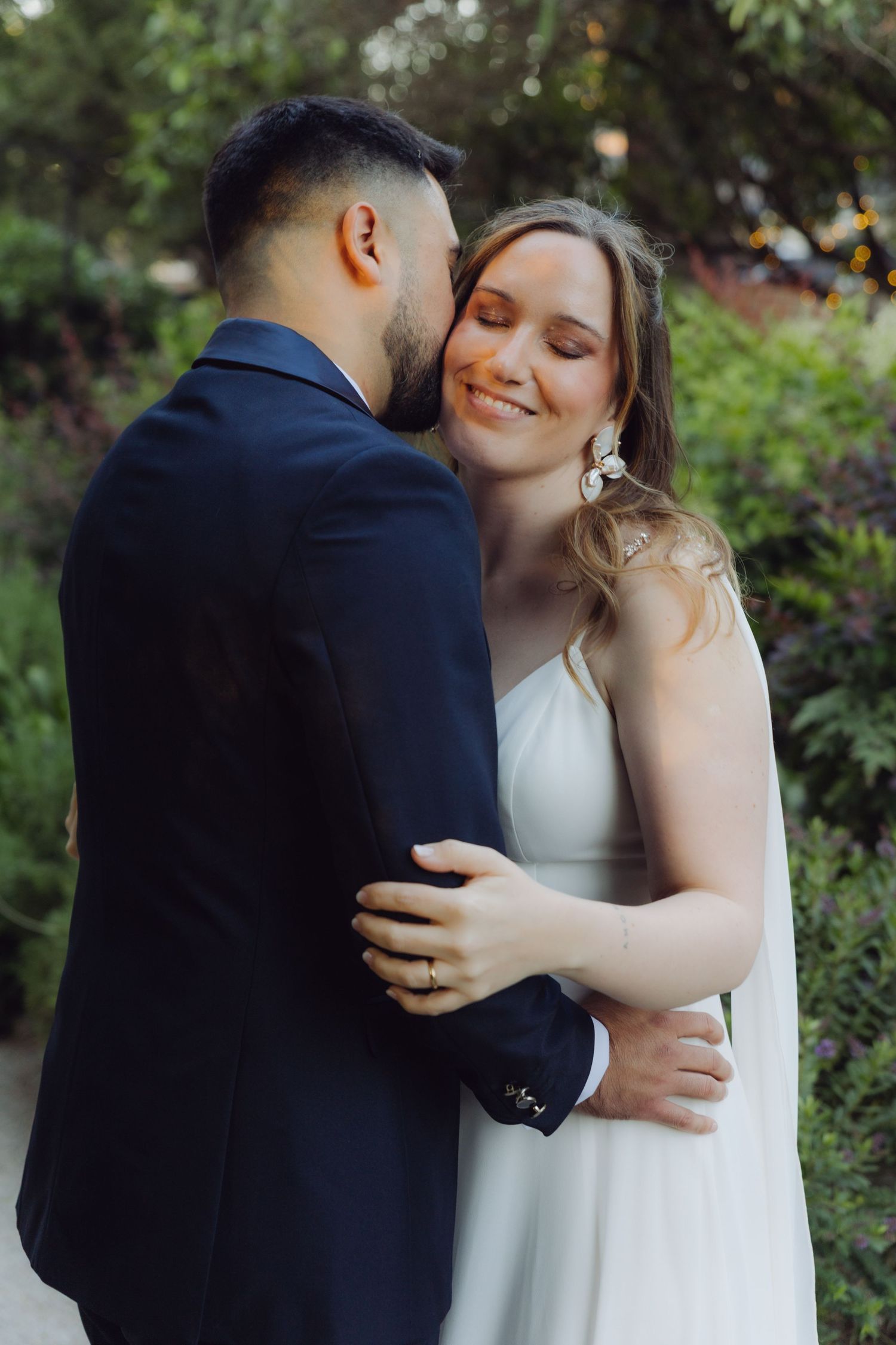 Bride smiles softly as groom kisses her cheek while they embrace in a garden.