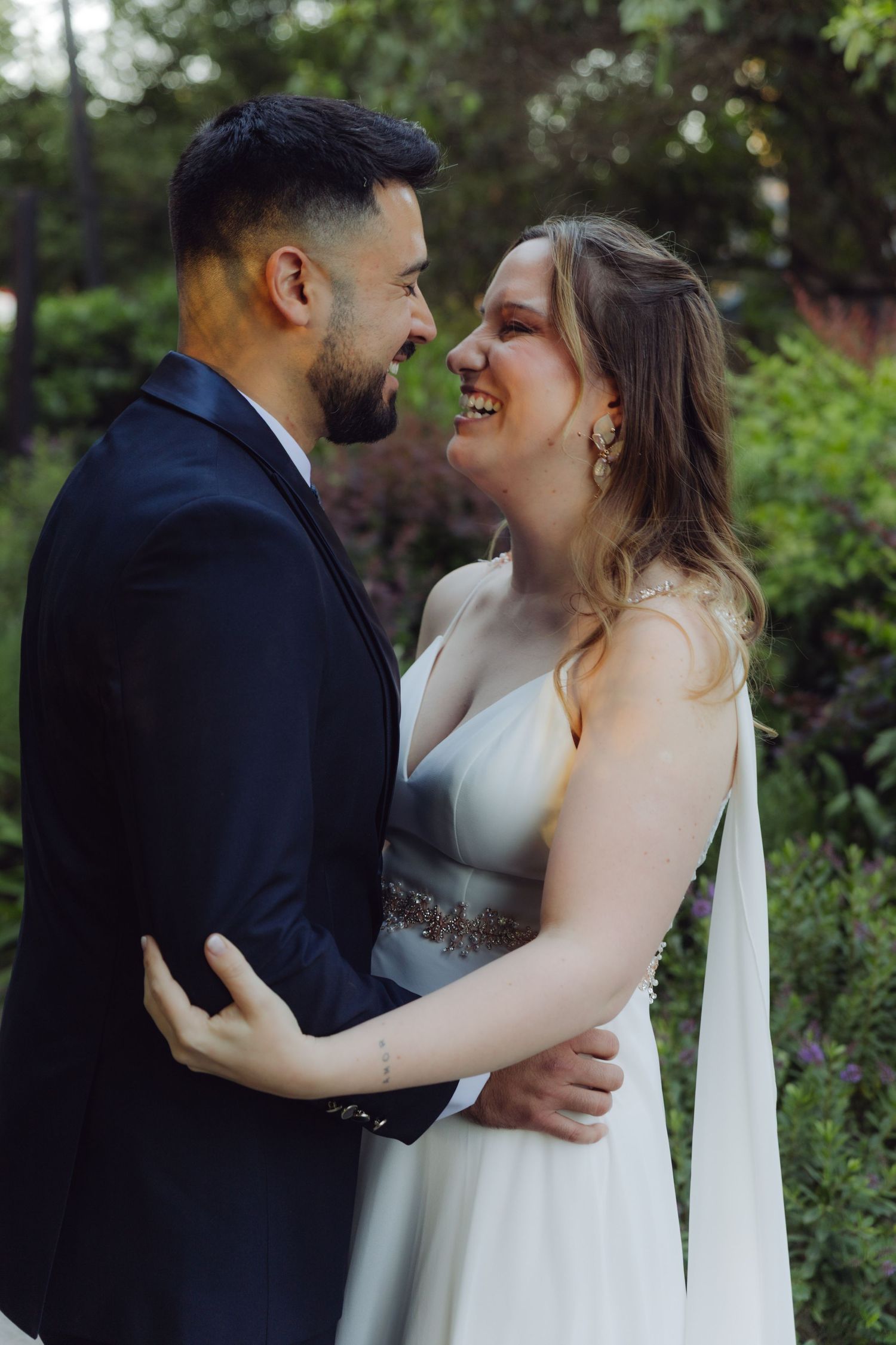 Bride and groom laugh together while embracing in a lush outdoor garden setting.