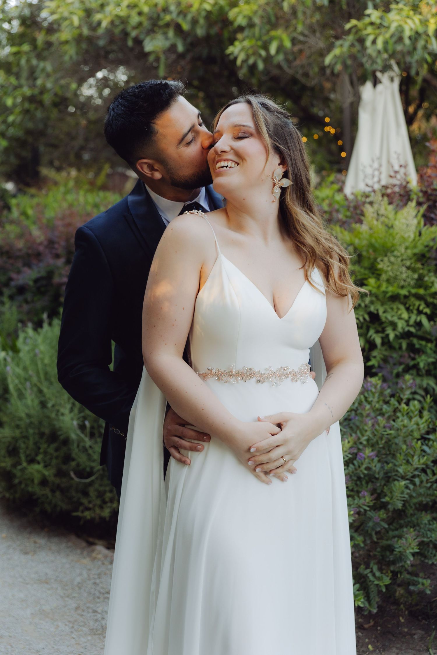 Groom kisses smiling bride on the cheek while embracing her in a garden setting.