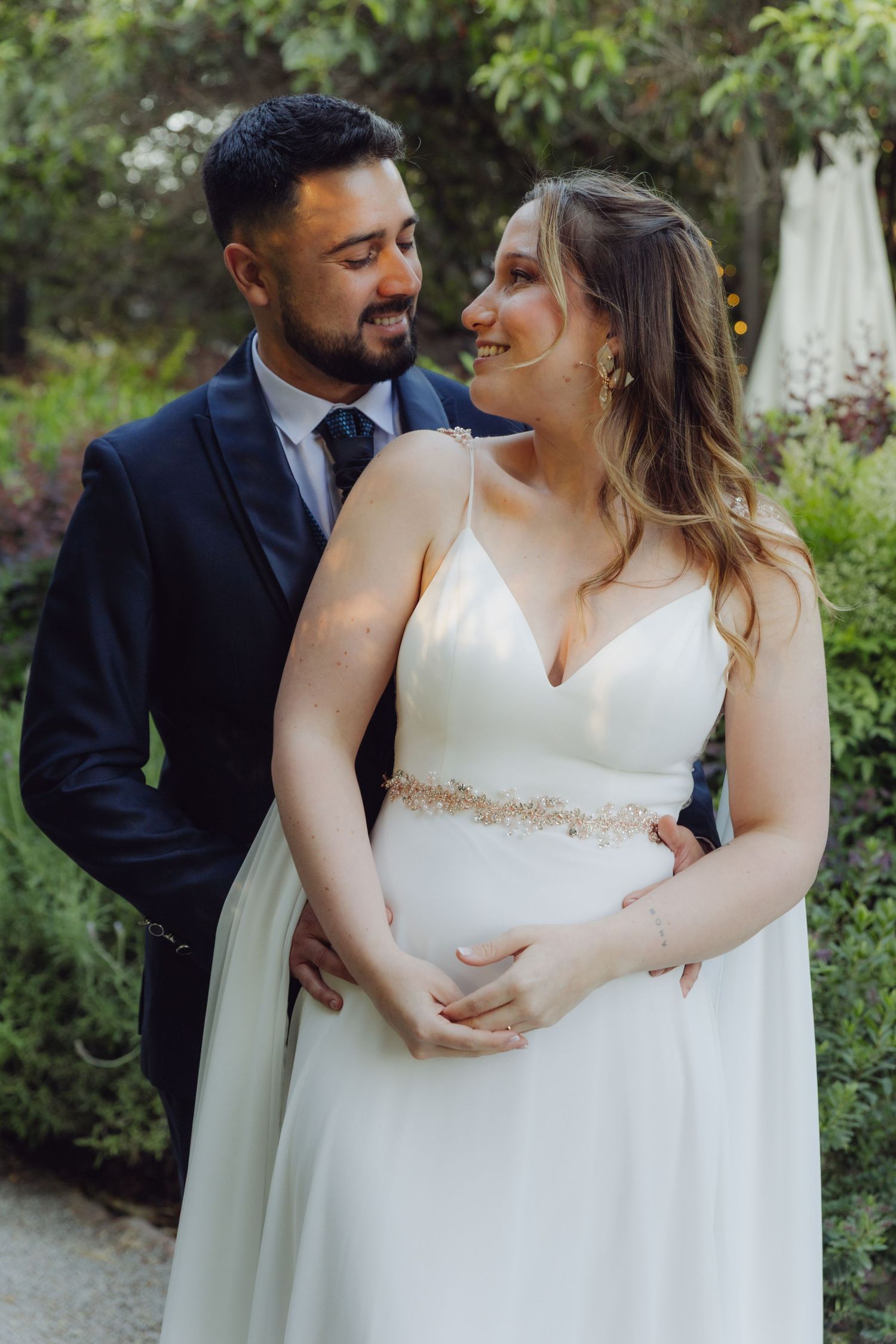 Smiling bride and groom embrace and gaze at each other in an outdoor garden.