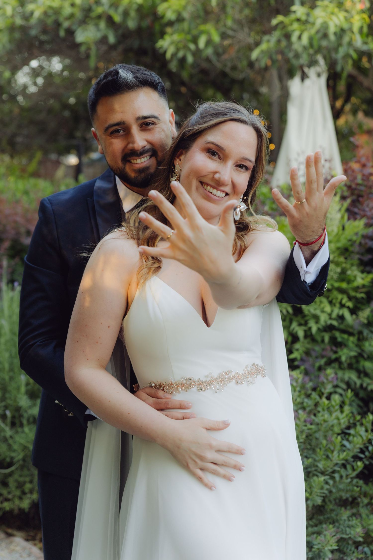 Happy bride and groom show off their wedding rings while posing in a garden.