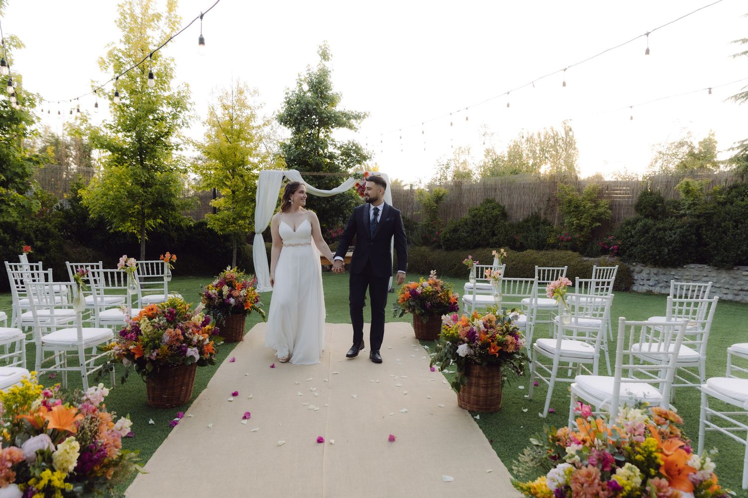 Bride and groom walk down outdoor wedding aisle lined with colorful floral arrangements and white chiavari chairs.