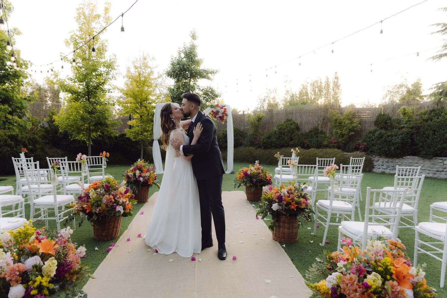 Newlyweds share first kiss at outdoor wedding ceremony surrounded by vibrant floral arrangements and white chairs.