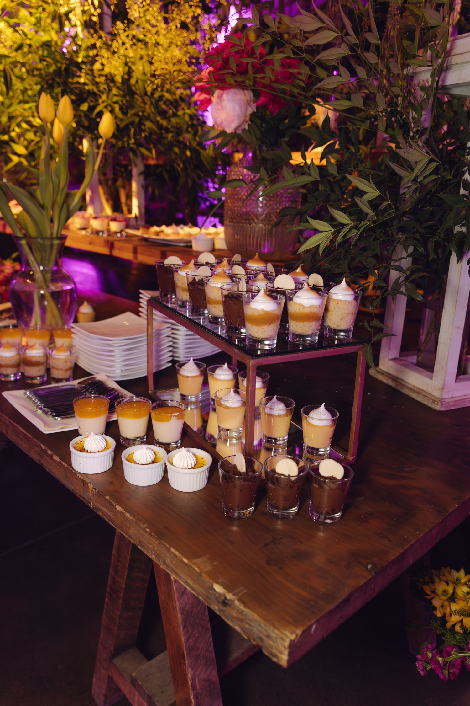 Rustic wooden table wedding dessert bar with assorted mousse cups and sweet treats amid lush floral decor.