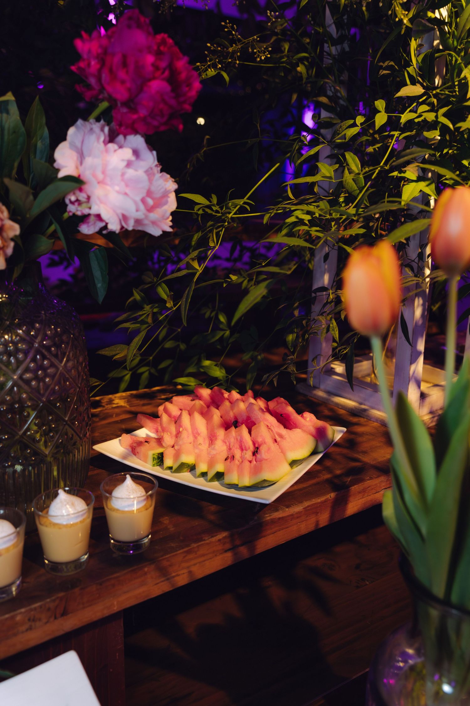 Wedding reception dessert table featuring sliced watermelon platter surrounded by colorful flowers and candles.