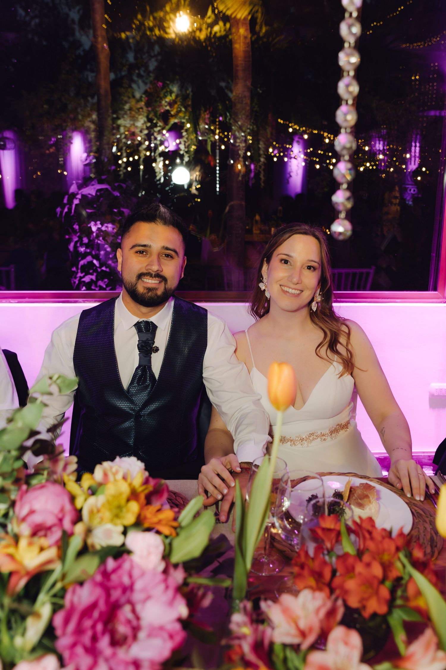 Smiling bride and groom seated at sweetheart table adorned with colorful flowers at evening wedding reception.