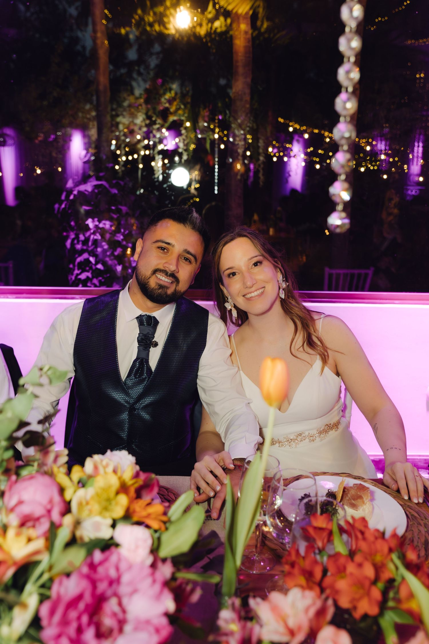 Happy newlyweds pose together at floral-decorated sweetheart table under purple-lit outdoor wedding reception.