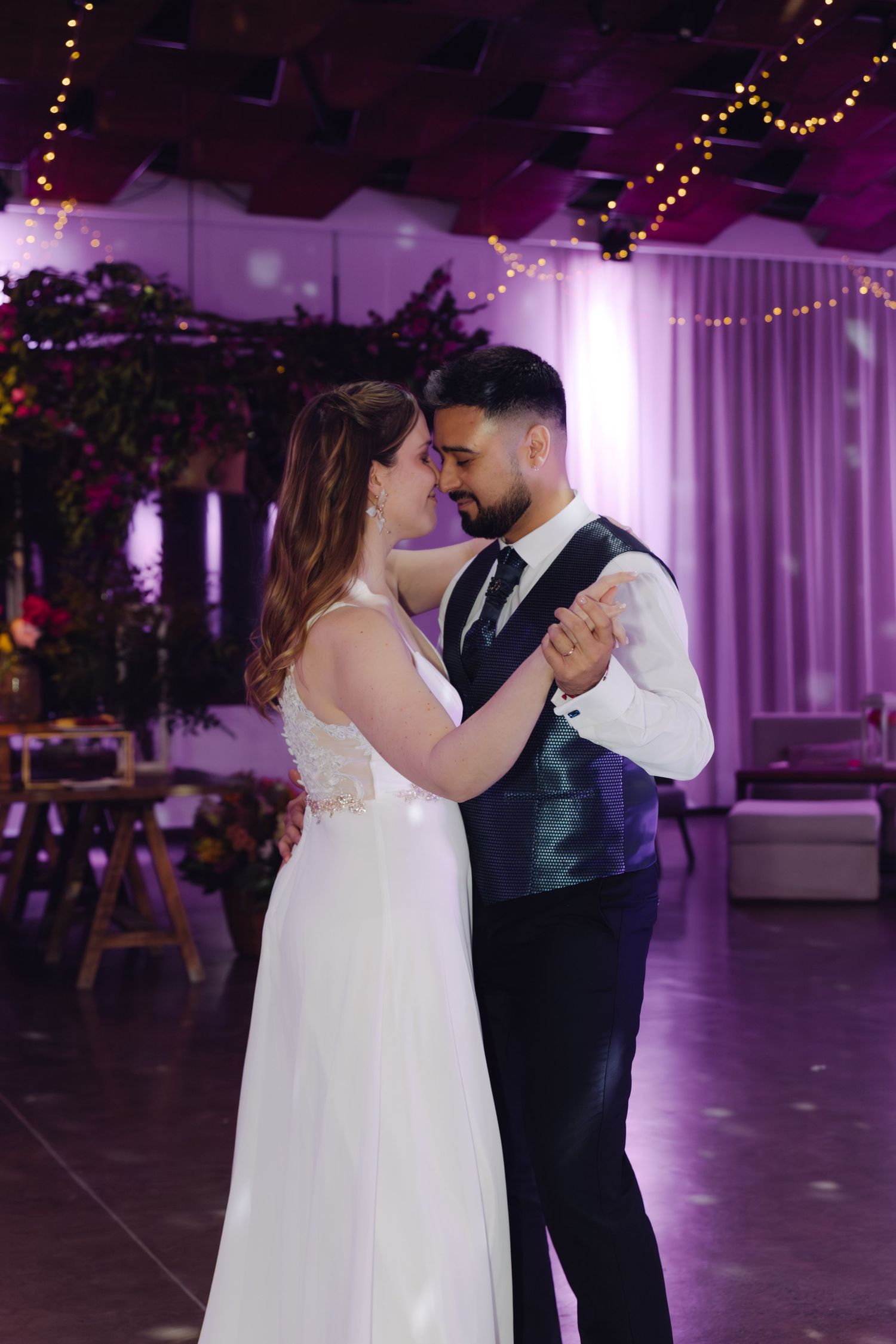 Bride and groom share intimate first dance in purple-lit wedding reception ballroom with string lights.