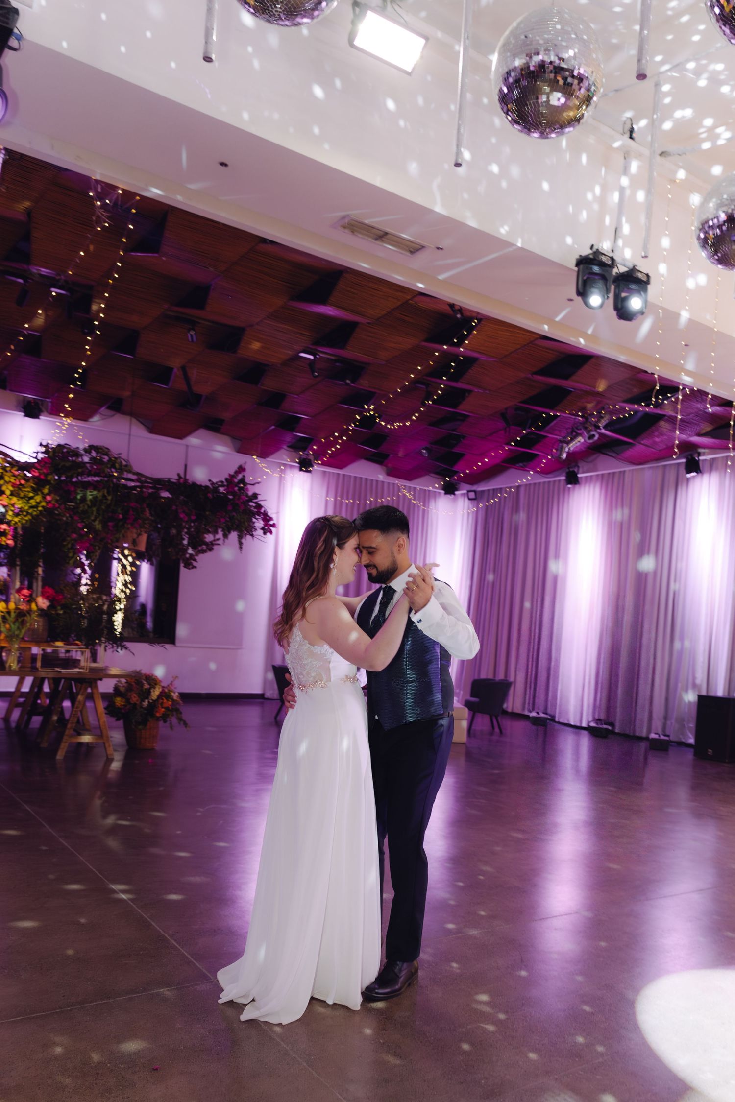 Newlyweds share romantic first dance under disco balls and string lights in purple-lit wedding reception hall.