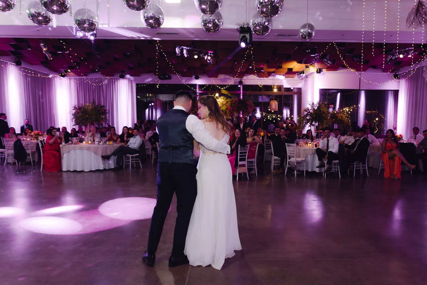 Bride and groom share their first dance in an elegant wedding reception hall with disco balls and purple lighting.
