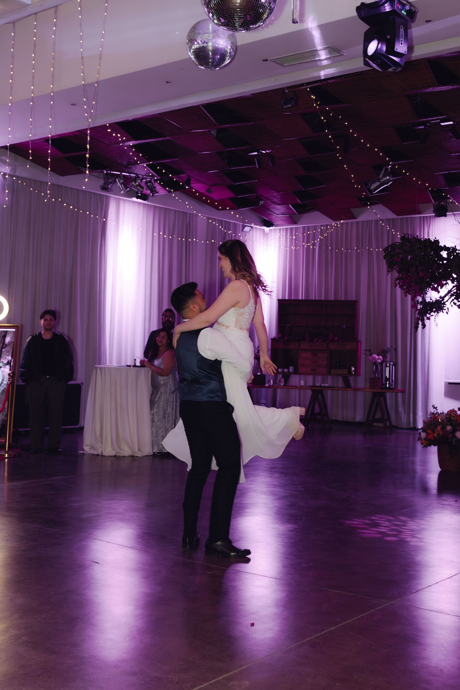 Groom lifts bride during their first dance in a beautifully decorated wedding reception hall with string lights.
