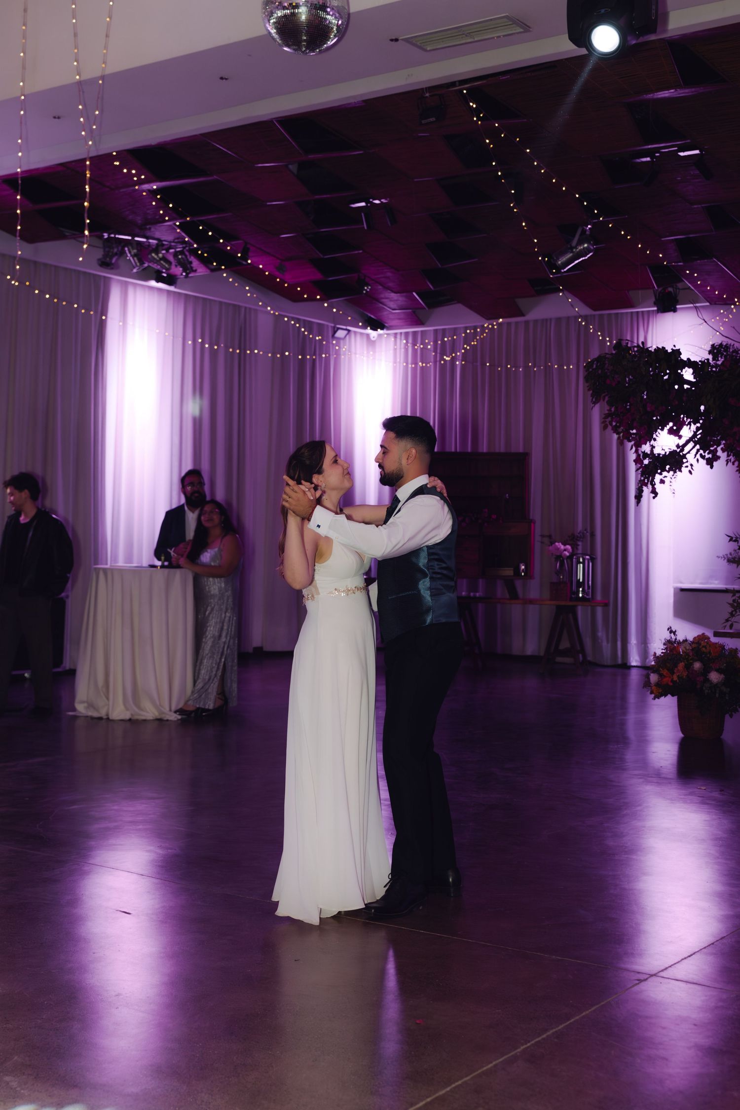 Newlyweds share an intimate first dance moment gazing at each other in a purple-lit wedding reception venue.