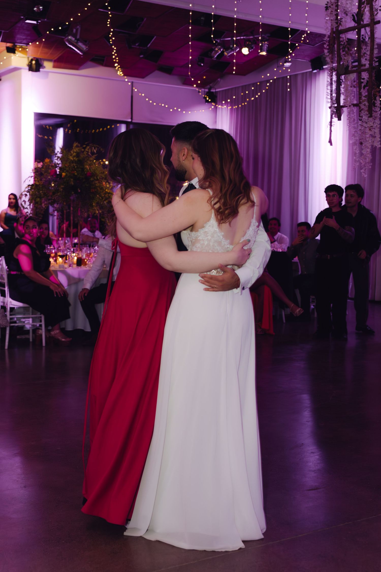Bride shares an emotional dance embrace with a woman in a red gown at a purple-lit wedding reception.