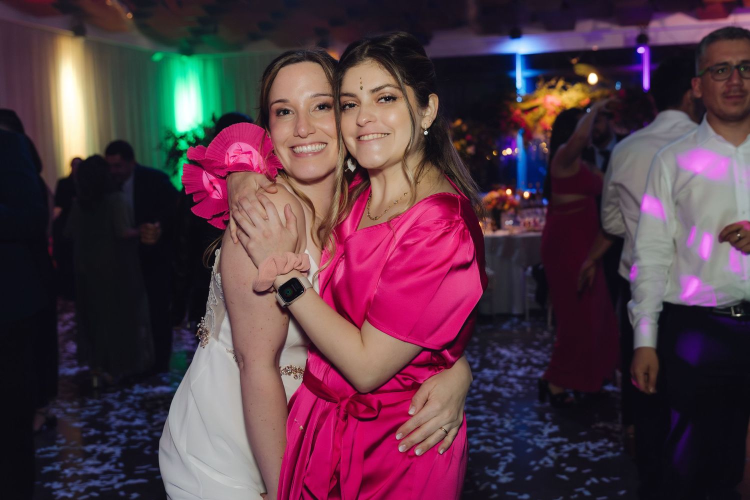 Bride in white gown hugs friend in pink dress at wedding reception celebration.