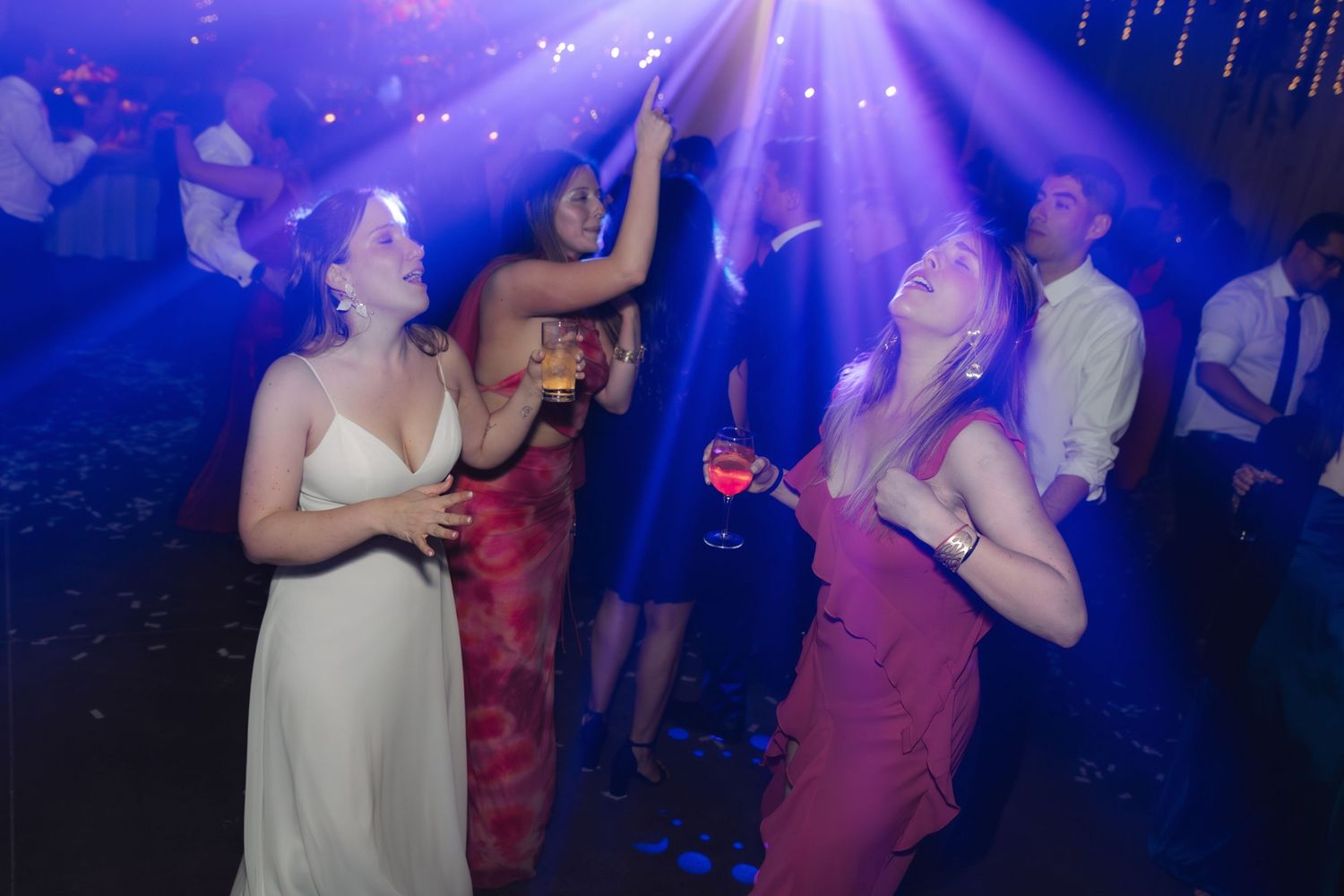 Women in formal gowns dancing under blue lights at a wedding reception.