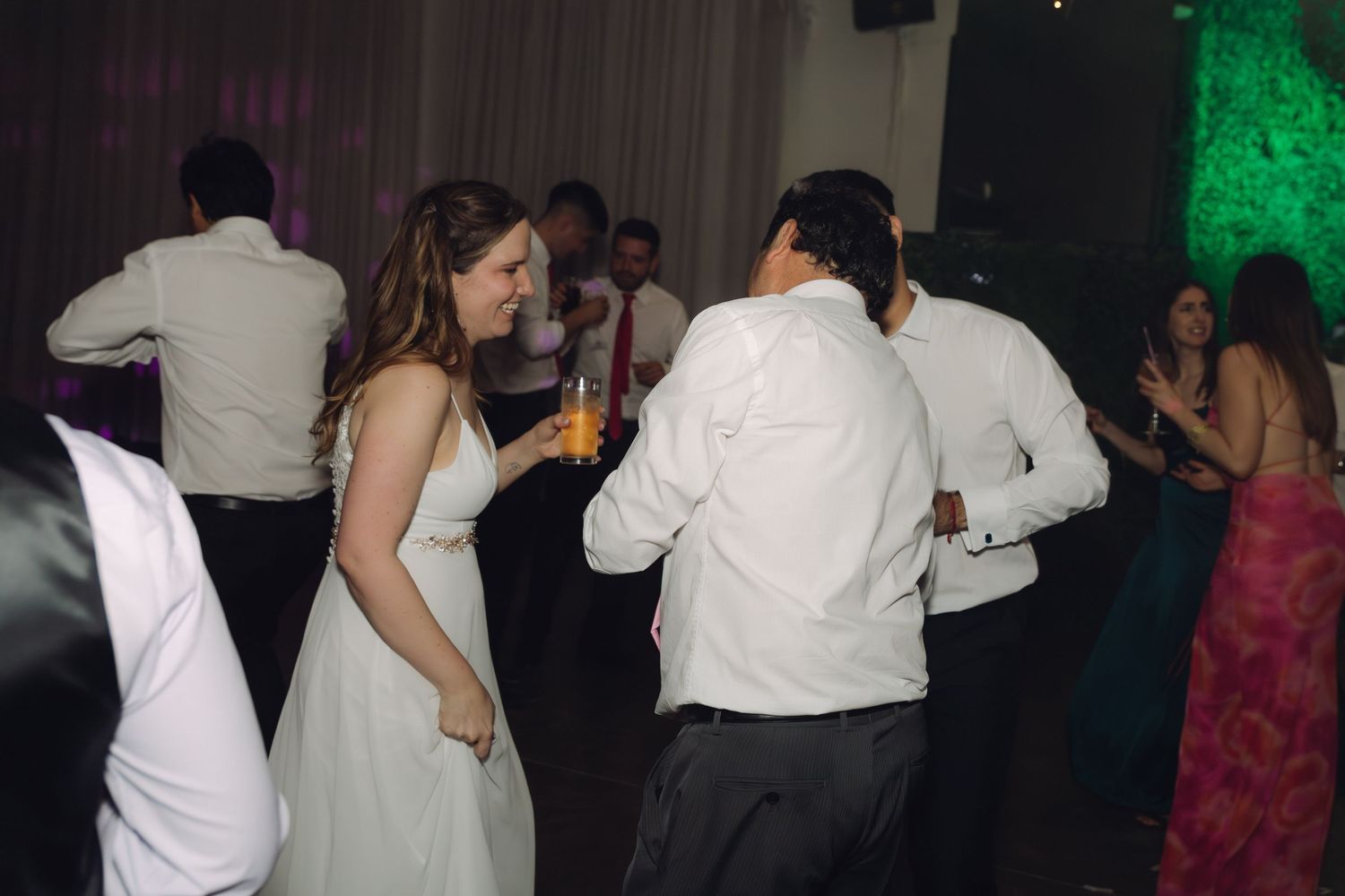 Bride in white gown dancing with guests at elegant wedding reception with colorful lights.